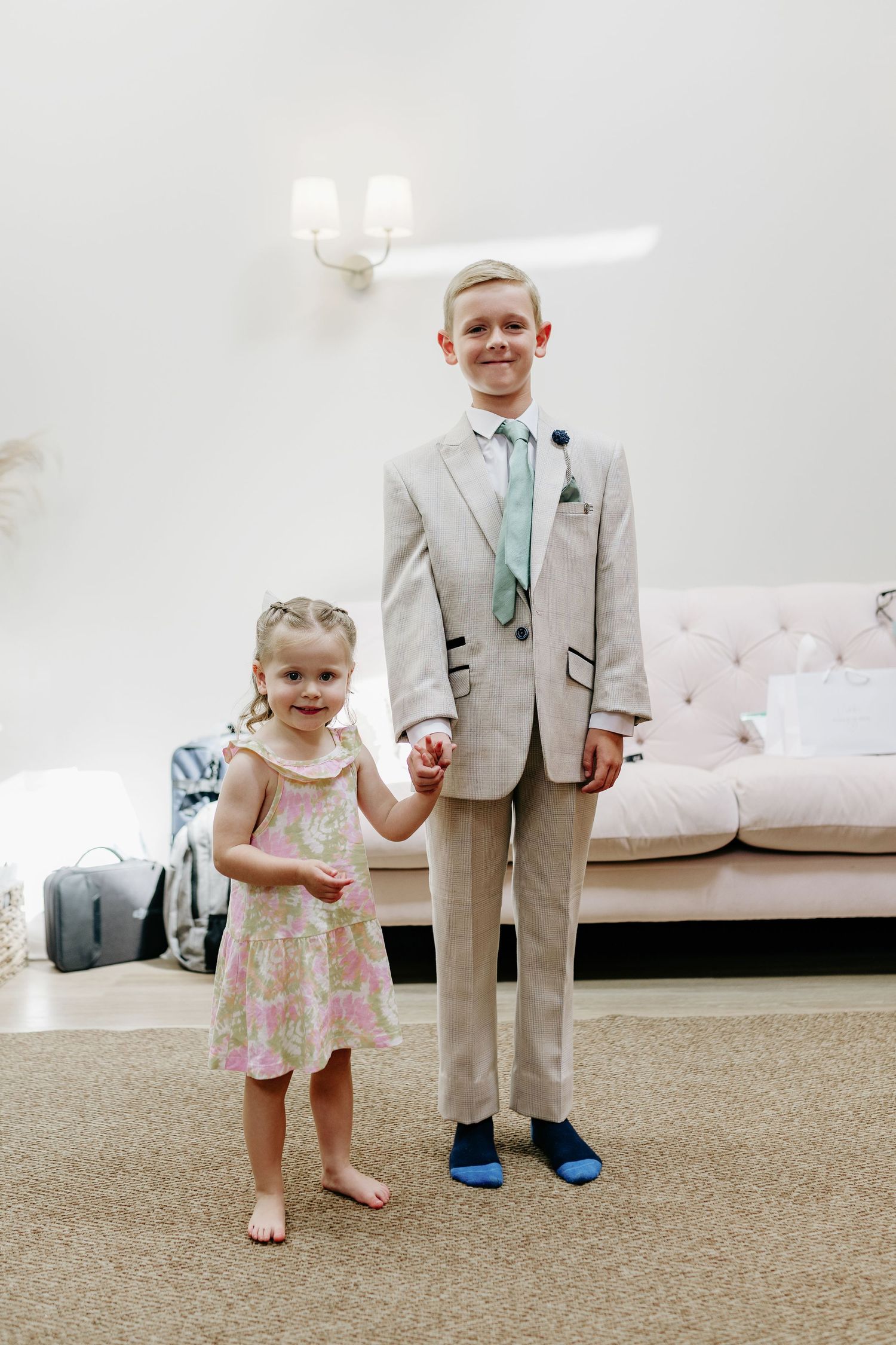 Two young children in wedding attire stand together on beige carpet in a bright indoor setting.