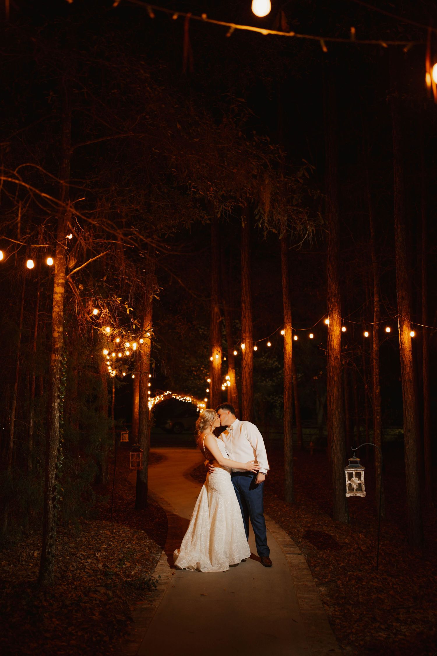 Romantic nighttime wedding portraits with string lights illuminating a wooded path.