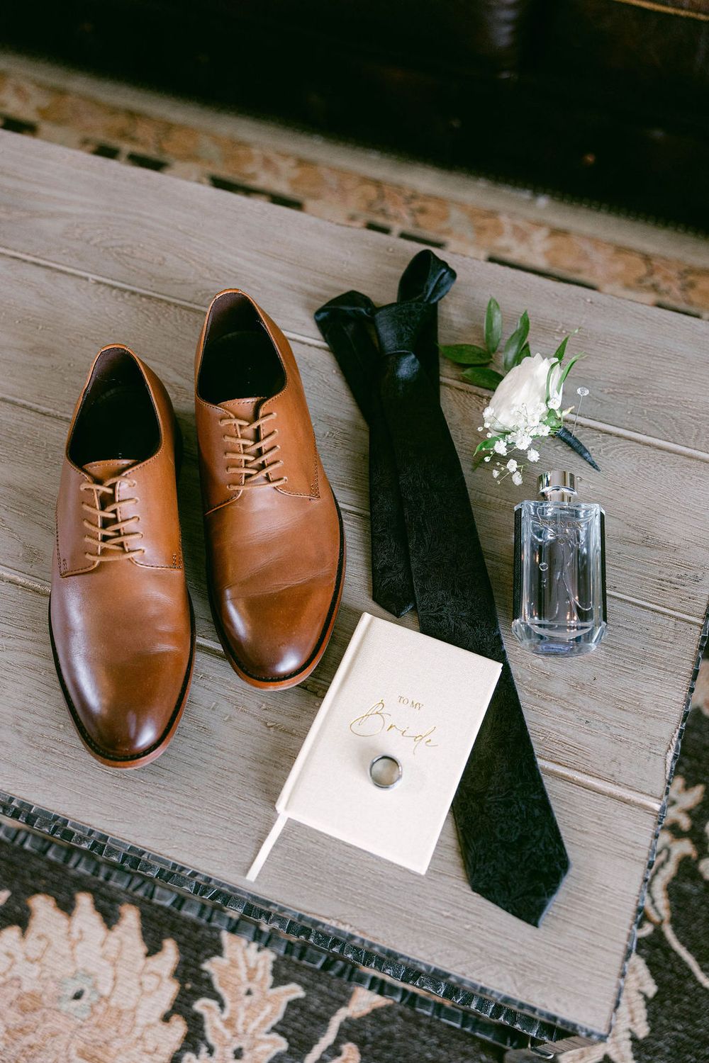 Classic brown leather dress shoes with a black tie and wedding invitation displayed on wooden surface next to small floral arrangement.