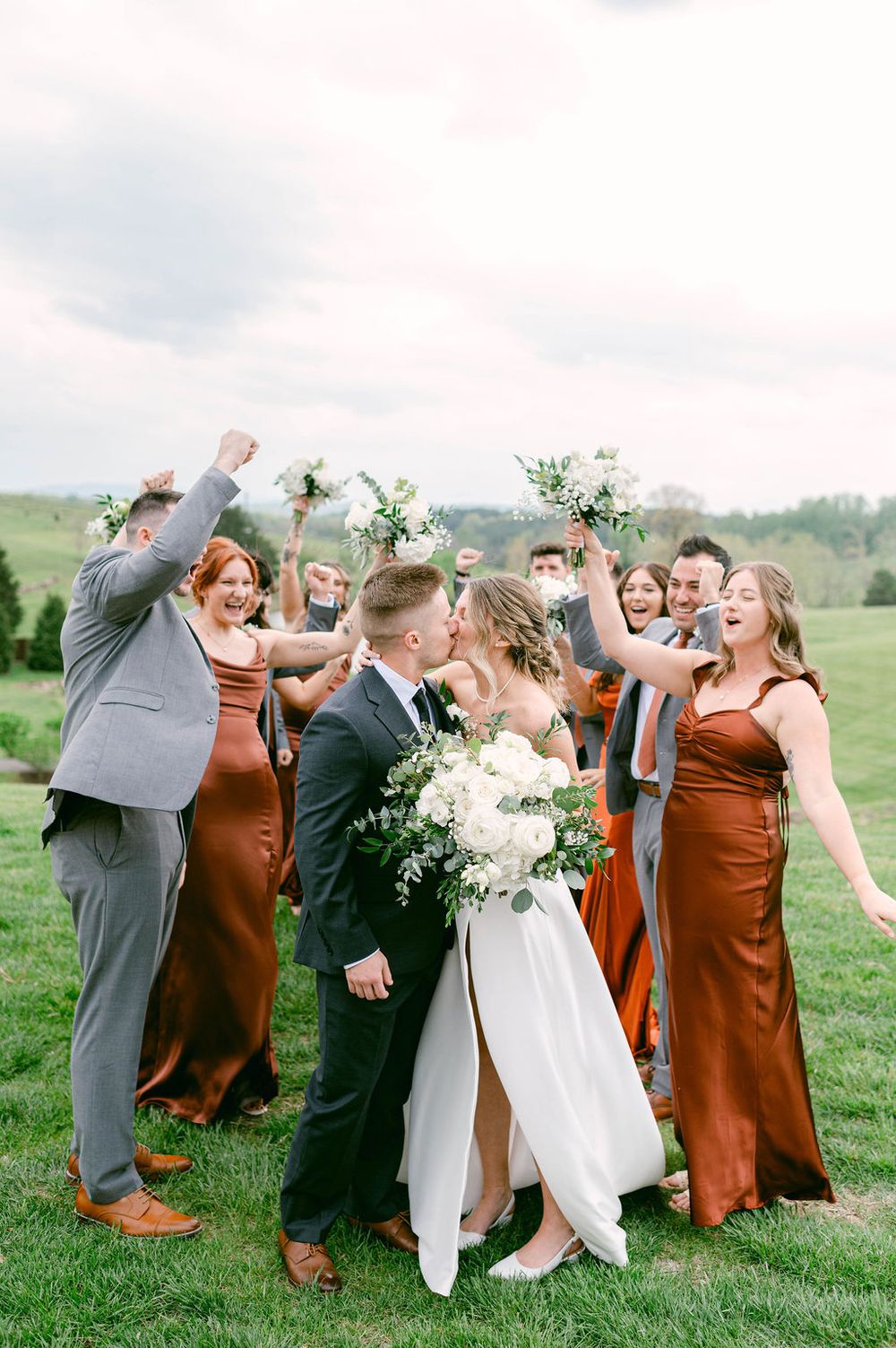 Wedding party wearing rust orange and gray attire poses playfully together on a grassy hillside during celebration.