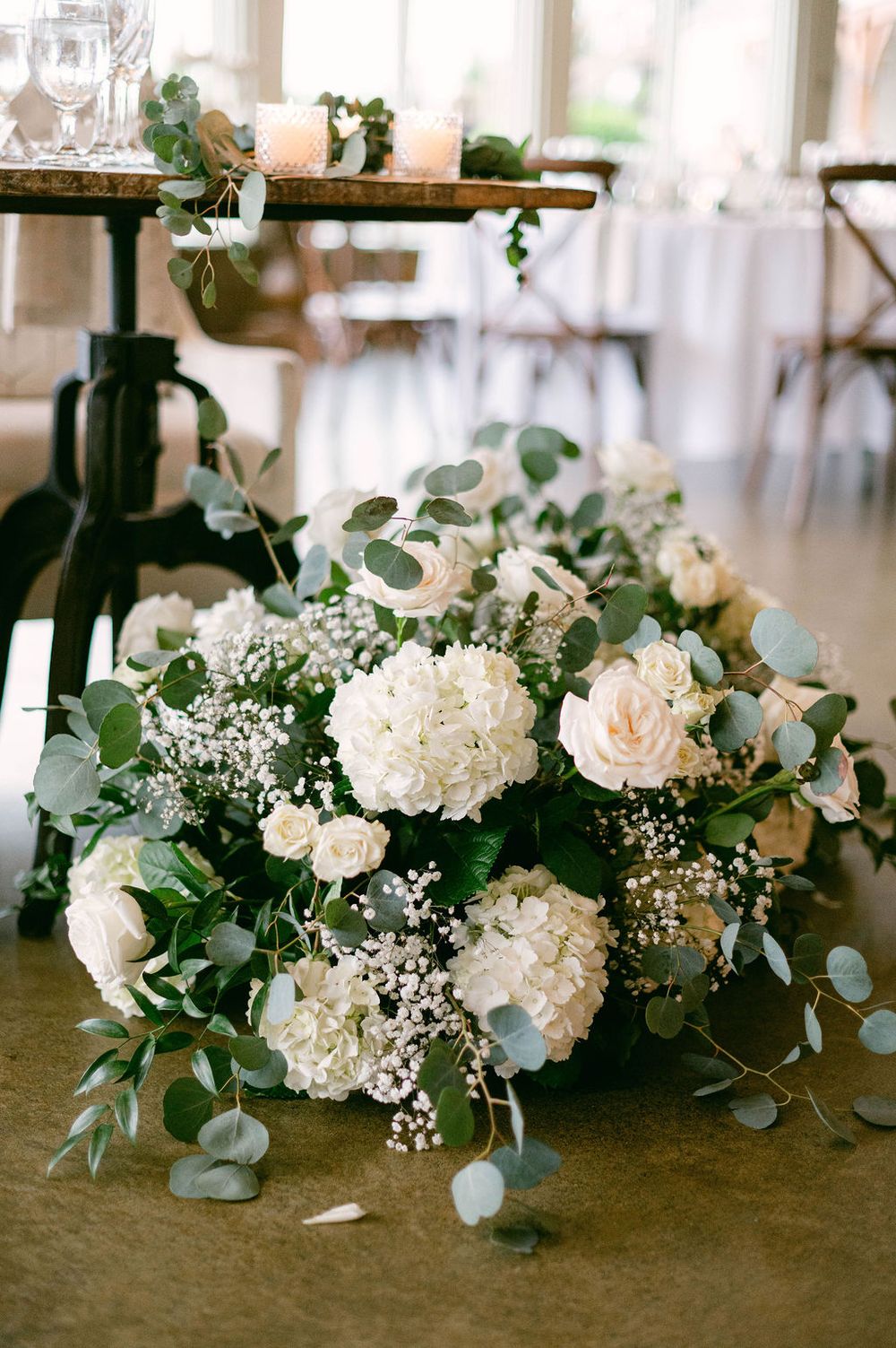 Elegant wedding floral arrangements with white roses, hydrangeas and eucalyptus leaves displayed on a wooden surface.