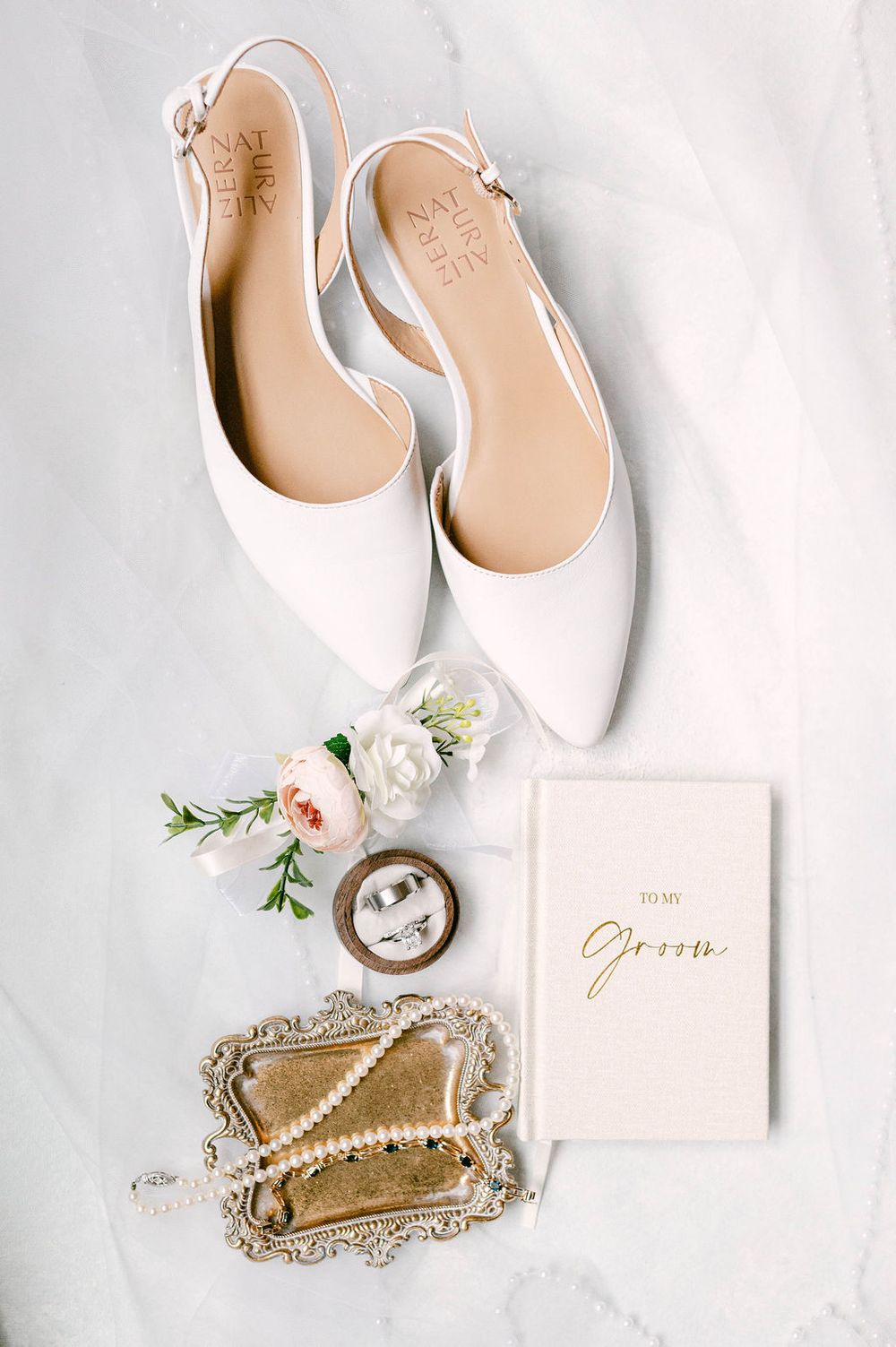 White bridal shoes, pearl necklace, and 'Groom' card arranged on an ornate gold tray against white background.