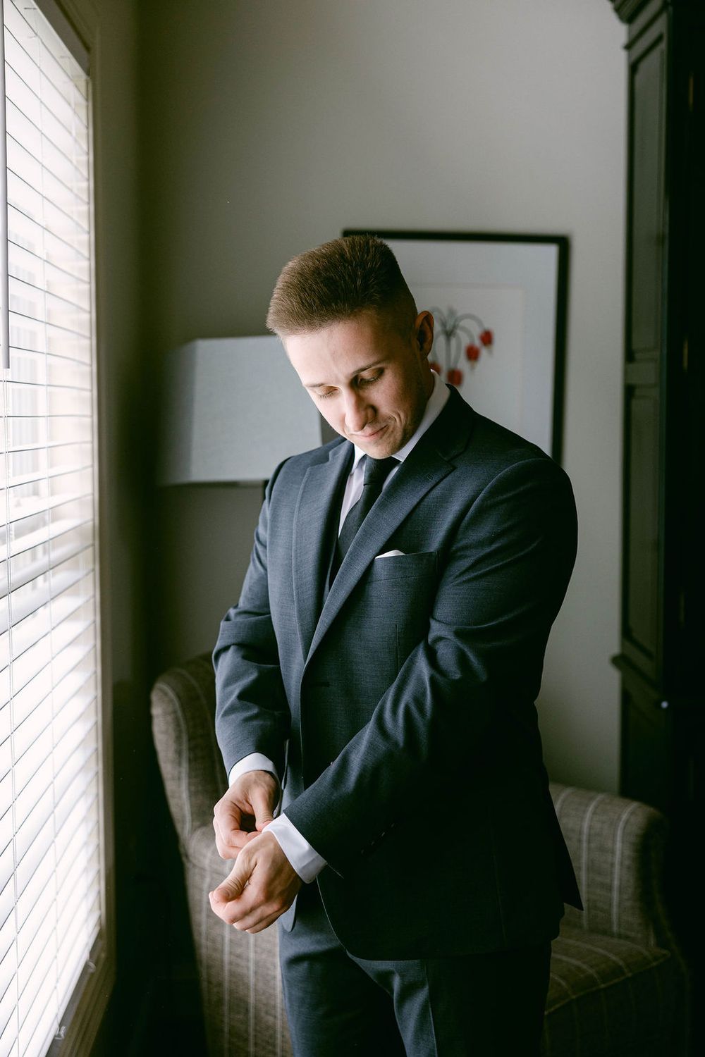 A person in a dark suit adjusts their cuffs while standing near a window in a dimly lit room.