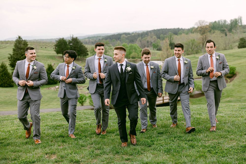Groomsmen in matching gray suits and red ties pose together on a grassy hill for wedding party photos.
