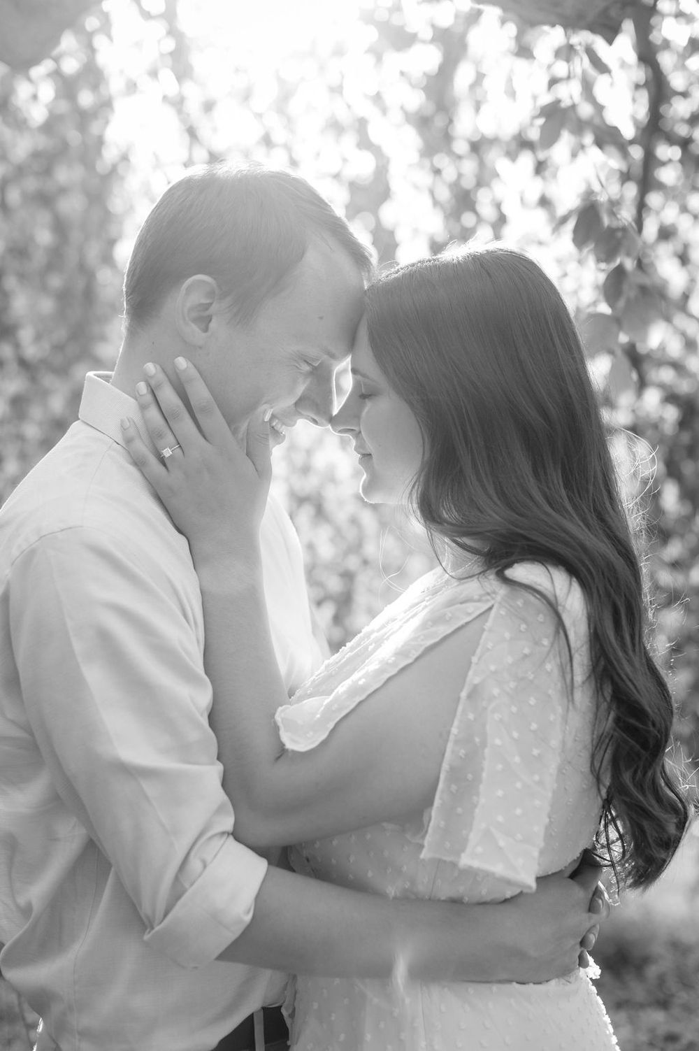 A romantic couple in white clothing shares a tender kiss outdoors amongst trees in black and white.