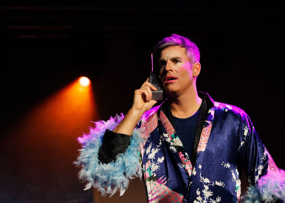 A performer in a navy floral shirt sings on stage under dramatic orange and purple lighting effects.