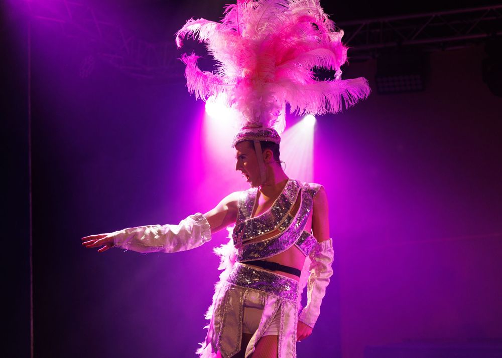 Performer in shimmering silver costume dances on stage illuminated by vibrant pink and purple theatrical lighting.