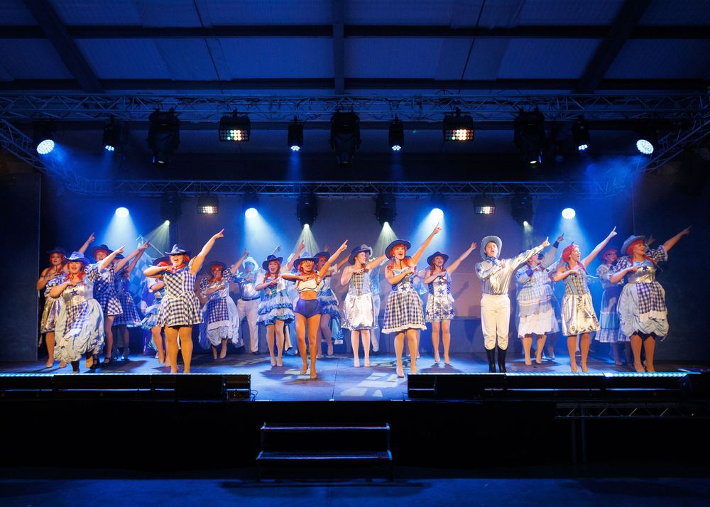 Performers in colorful costumes dance in formation on a stage illuminated by blue theatrical lighting.