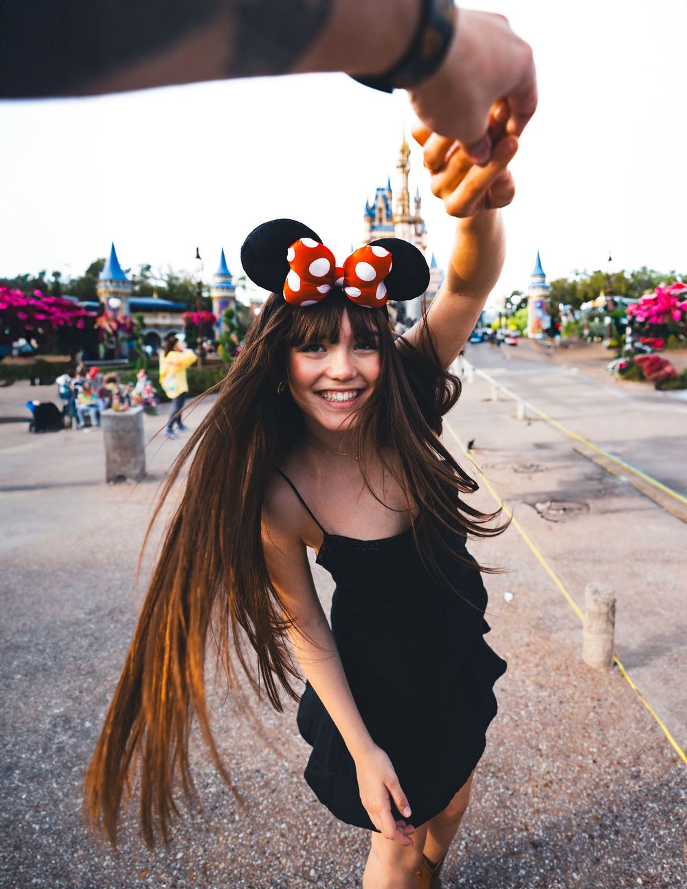 A person in a black top playfully twirls their long brown hair against a sunset sky in a parking lot.
