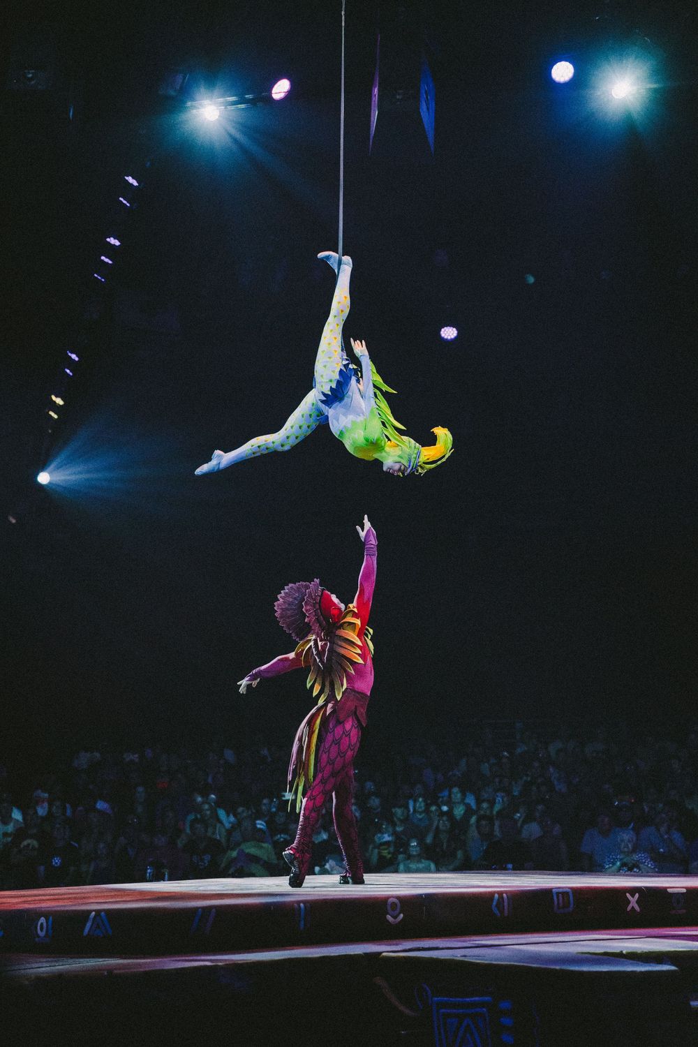 Acrobatic performers in colorful costumes execute dramatic aerial stunts on stage with blue lighting effects.