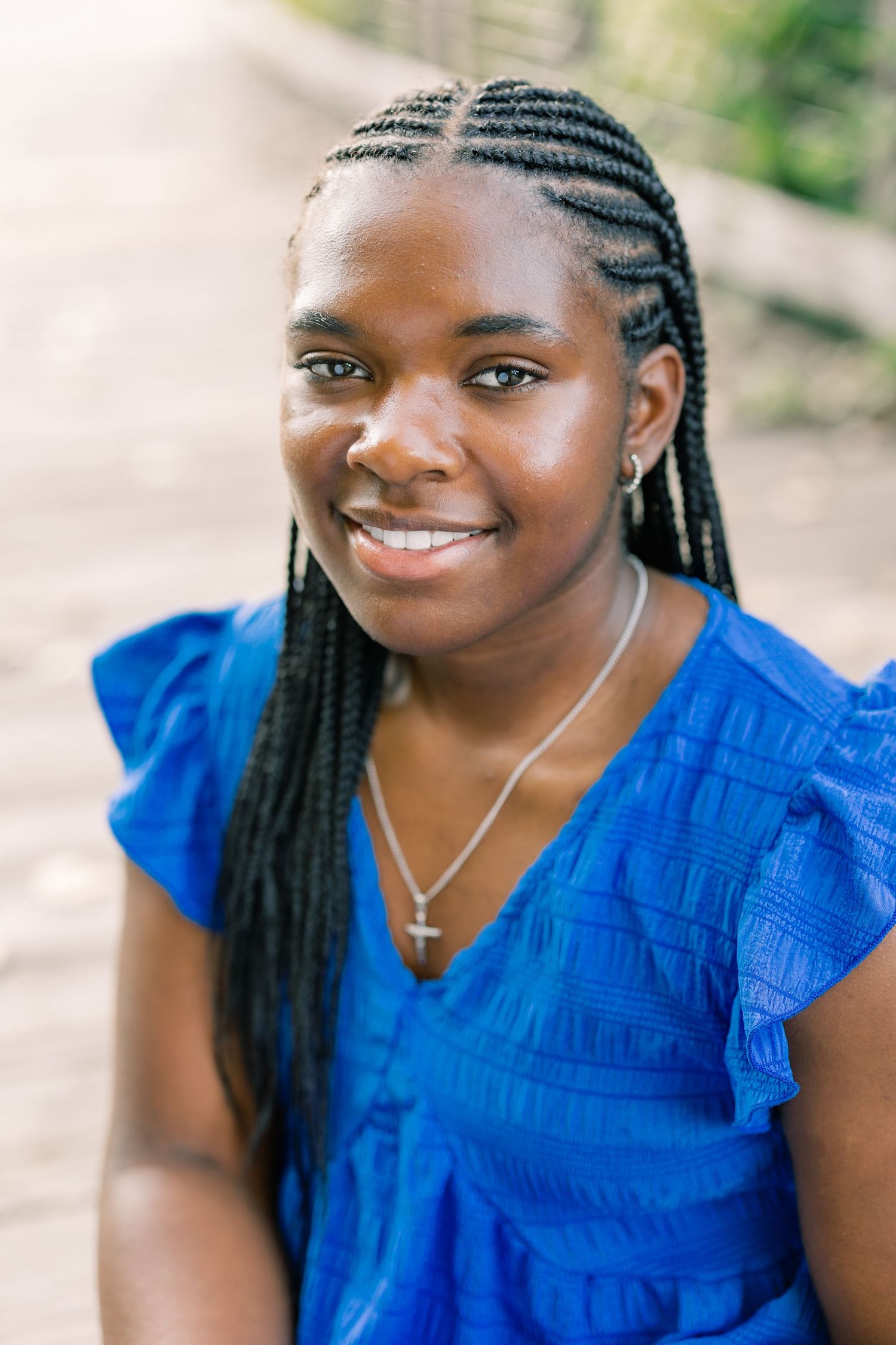 Person in a bright blue ruffled top smiles warmly while wearing cornrow braids and a cross necklace.