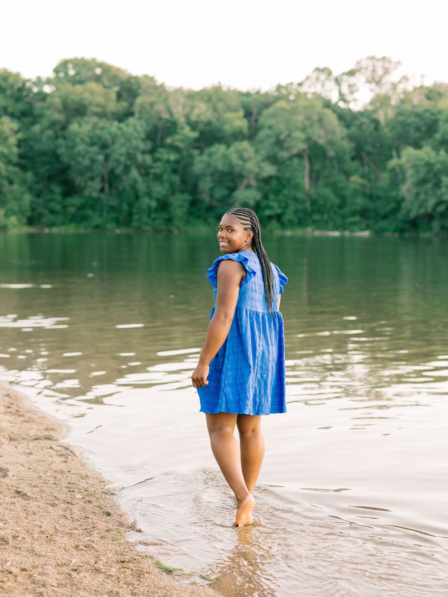A person in a bright blue summer dress walks barefoot along the shoreline of a serene lake surrounded by green forest.