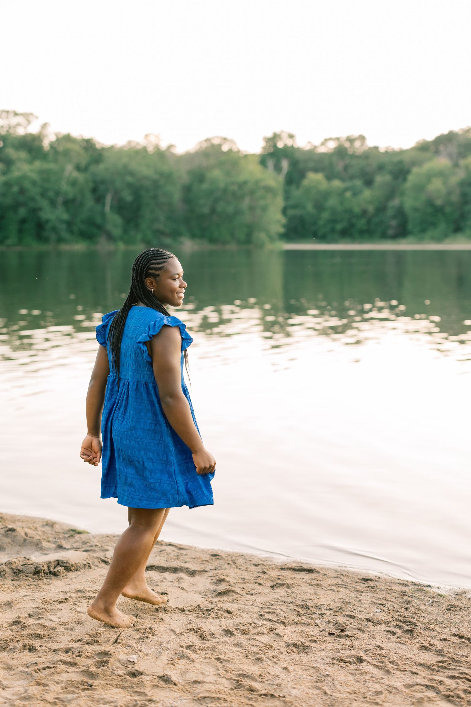 Person in a blue dress stands barefoot on a sandy beach beside a serene lake during golden hour.