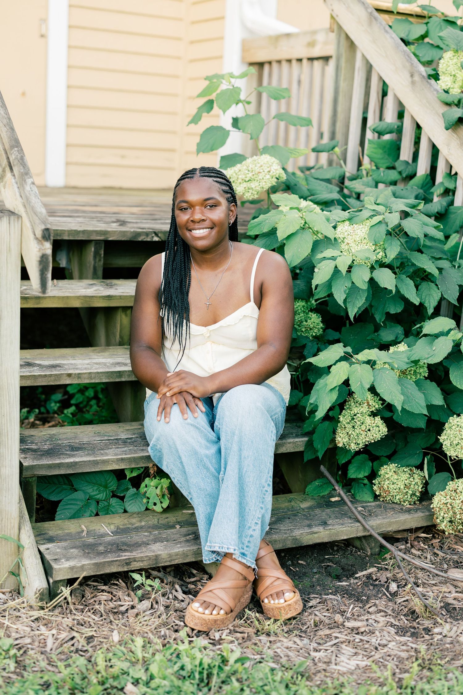Someone sits casually on wooden steps wearing a white tank top and light jeans near leafy green hydrangea plants.