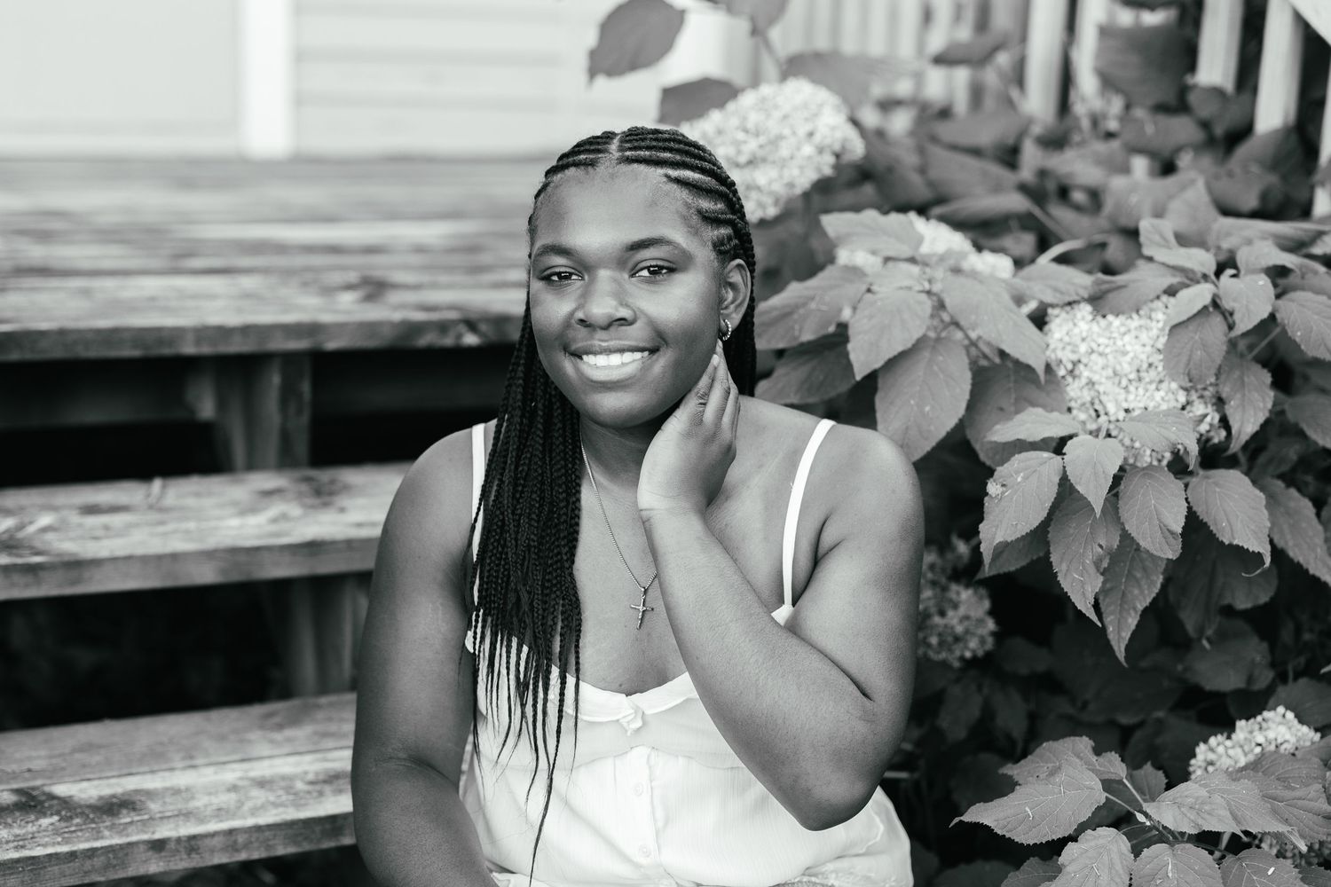 Black and white portrait of someone sitting on outdoor steps, smiling with hand on cheek near flowering hydrangea bushes.