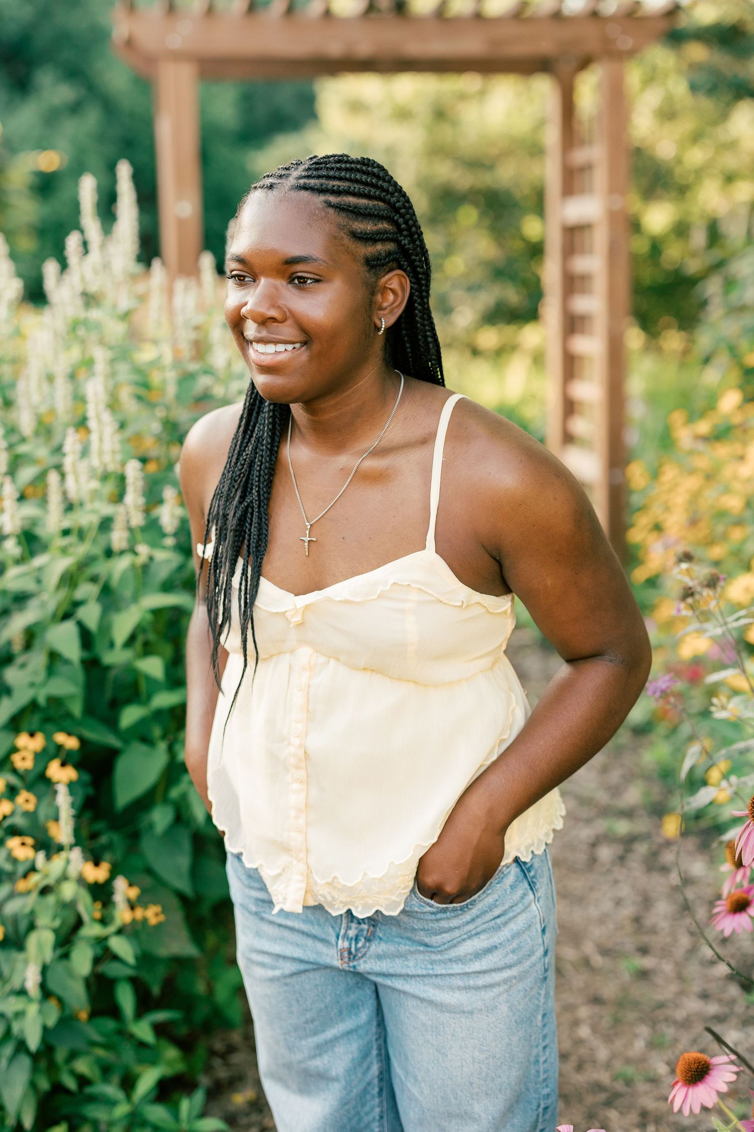 A person in a white camisole and light jeans poses in a summer garden with blooming flowers.