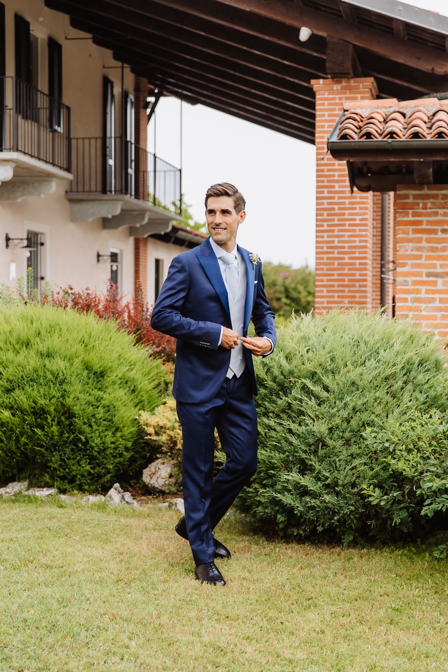 A person in a navy blue suit stands on a lawn with brick architecture and greenery in the background.