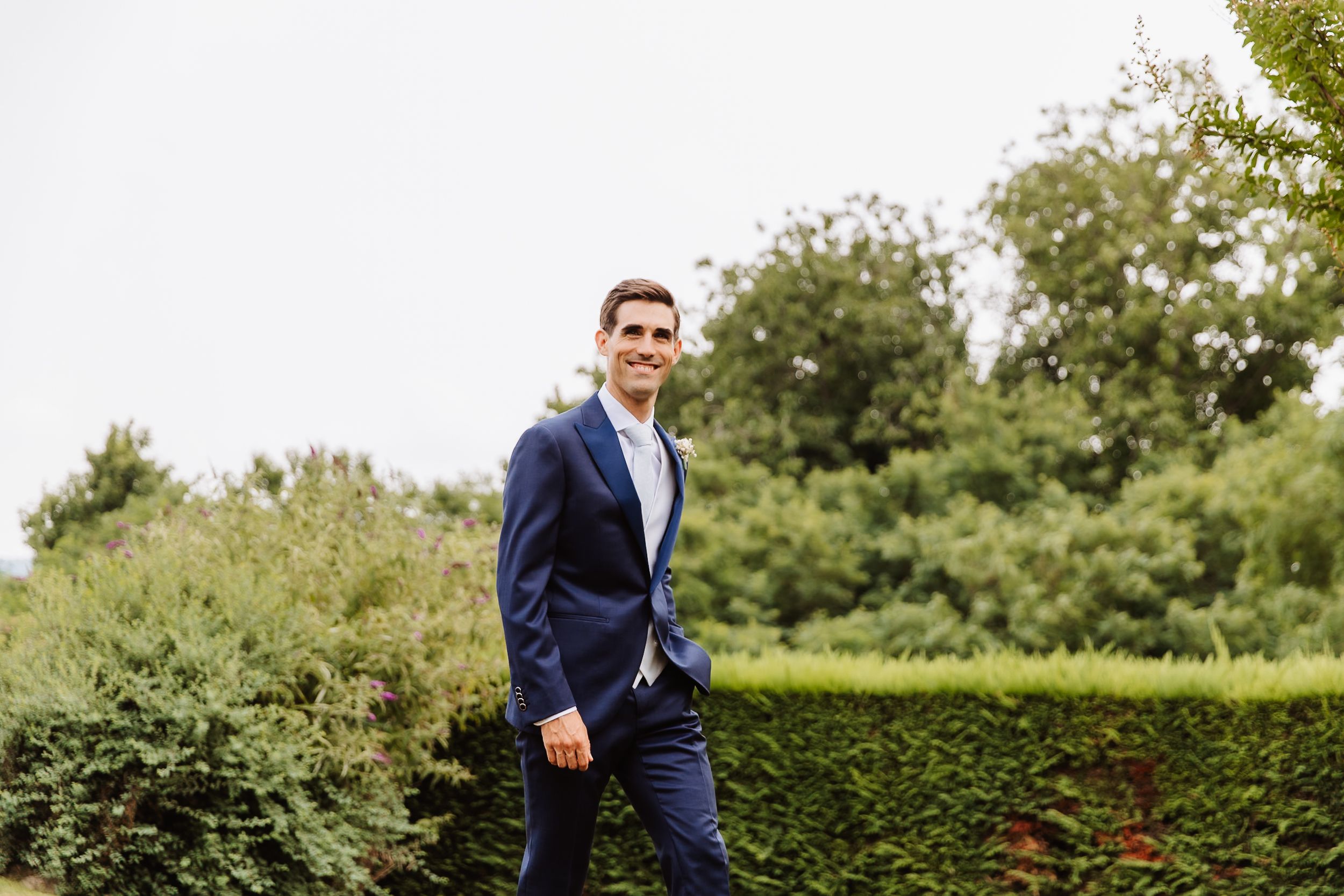 Person wearing a navy suit and blue shirt walks along a manicured hedge garden path on a sunny day.