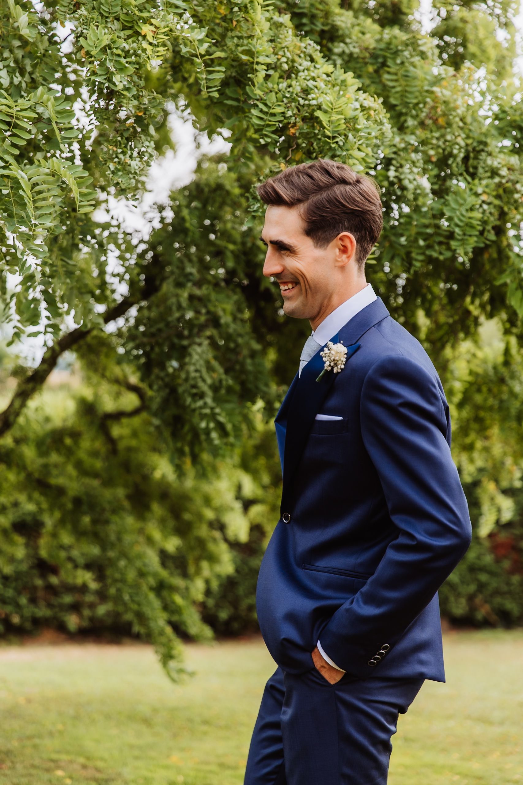 A figure wearing a navy blue suit stands in a lush outdoor setting surrounded by green leafy trees.