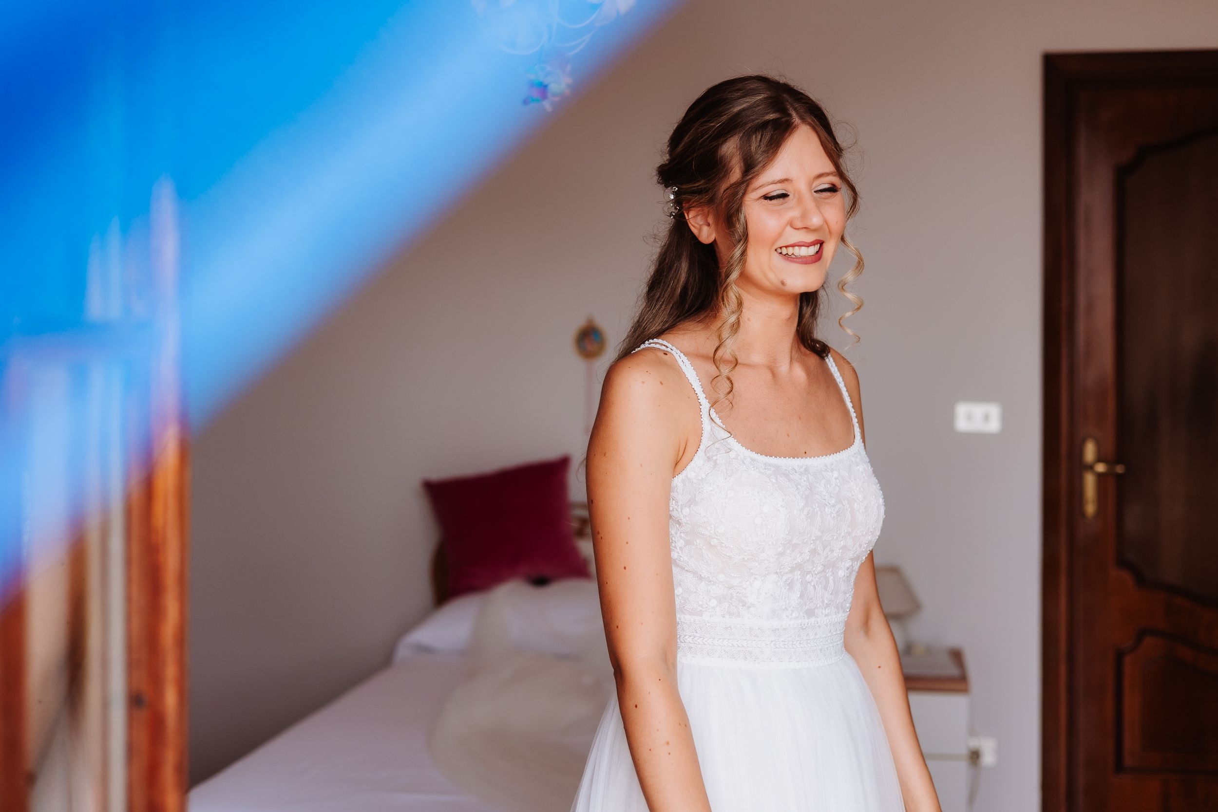 A serene bride in a flowing white lace dress smiles joyfully in a sunlit room during wedding preparations.