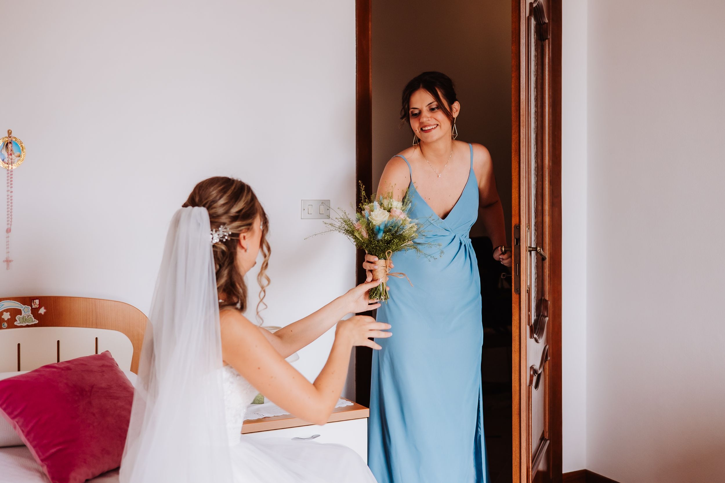 Two women in a bridal suite, one wearing a blue bridesmaid dress and another in a wedding gown holding a bouquet.