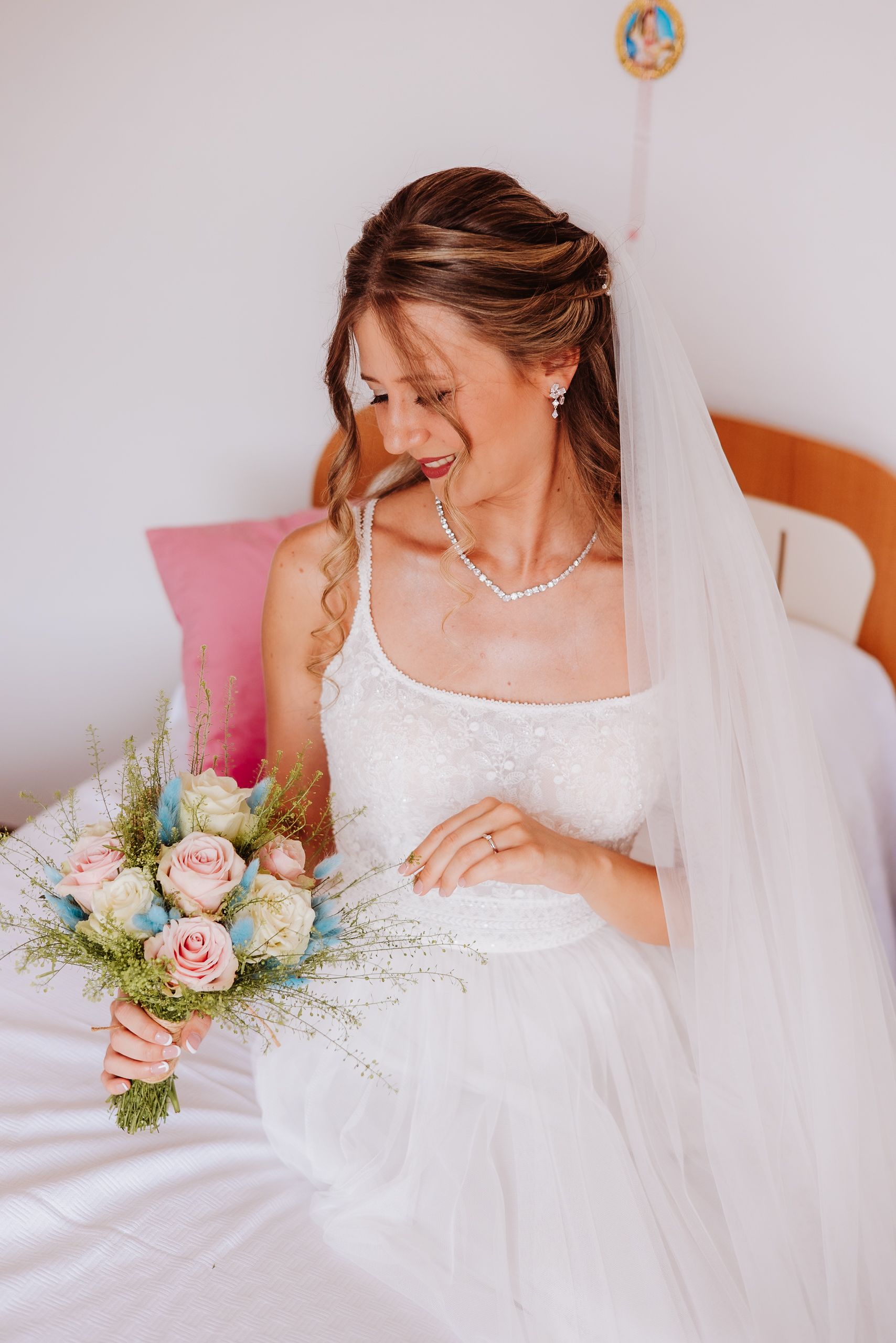 A bride in a white lace dress and veil sits on a bed holding a pink rose bouquet while smiling candidly.