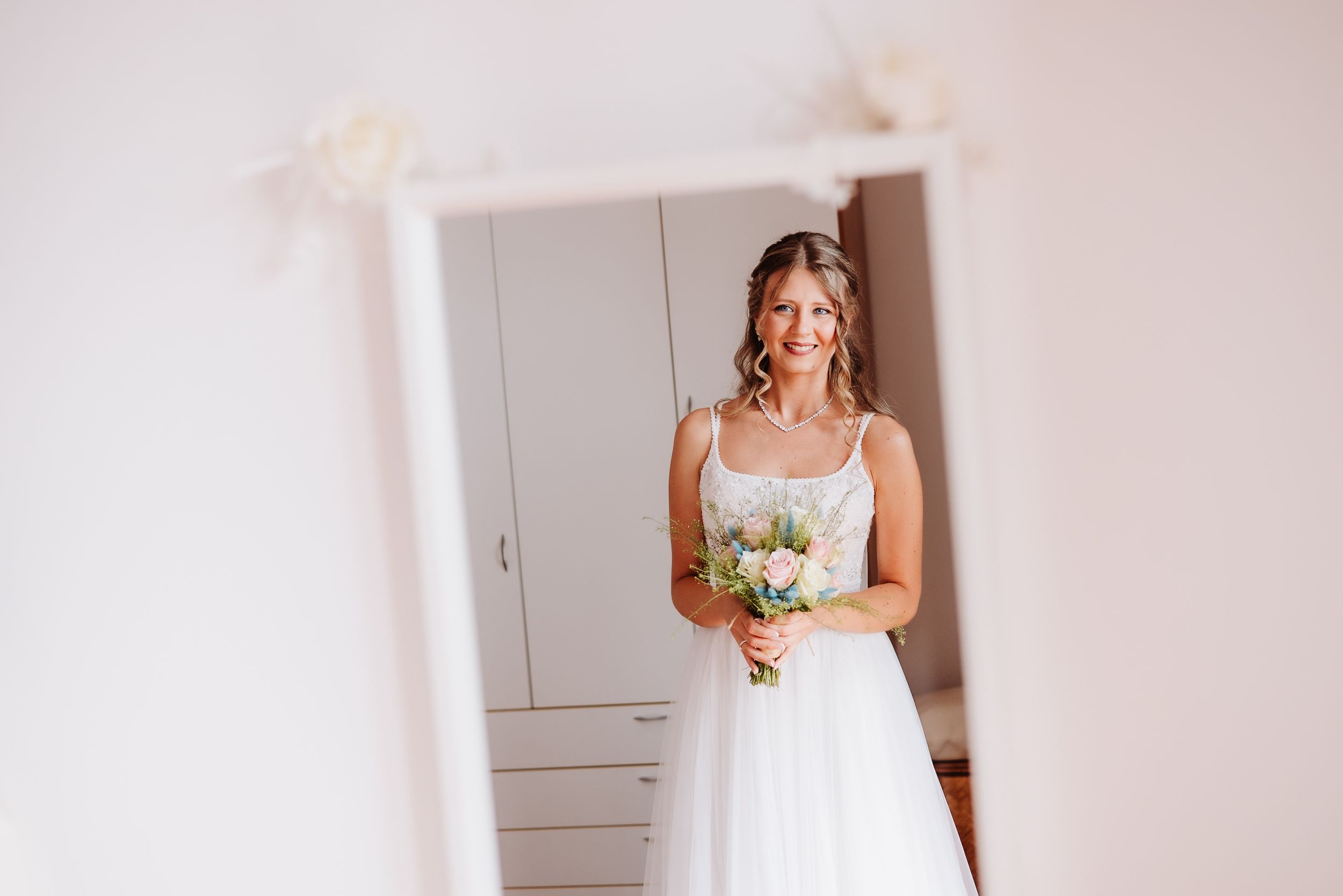 A bride in a white wedding dress holds a bouquet while standing next to a bright window with natural lighting.