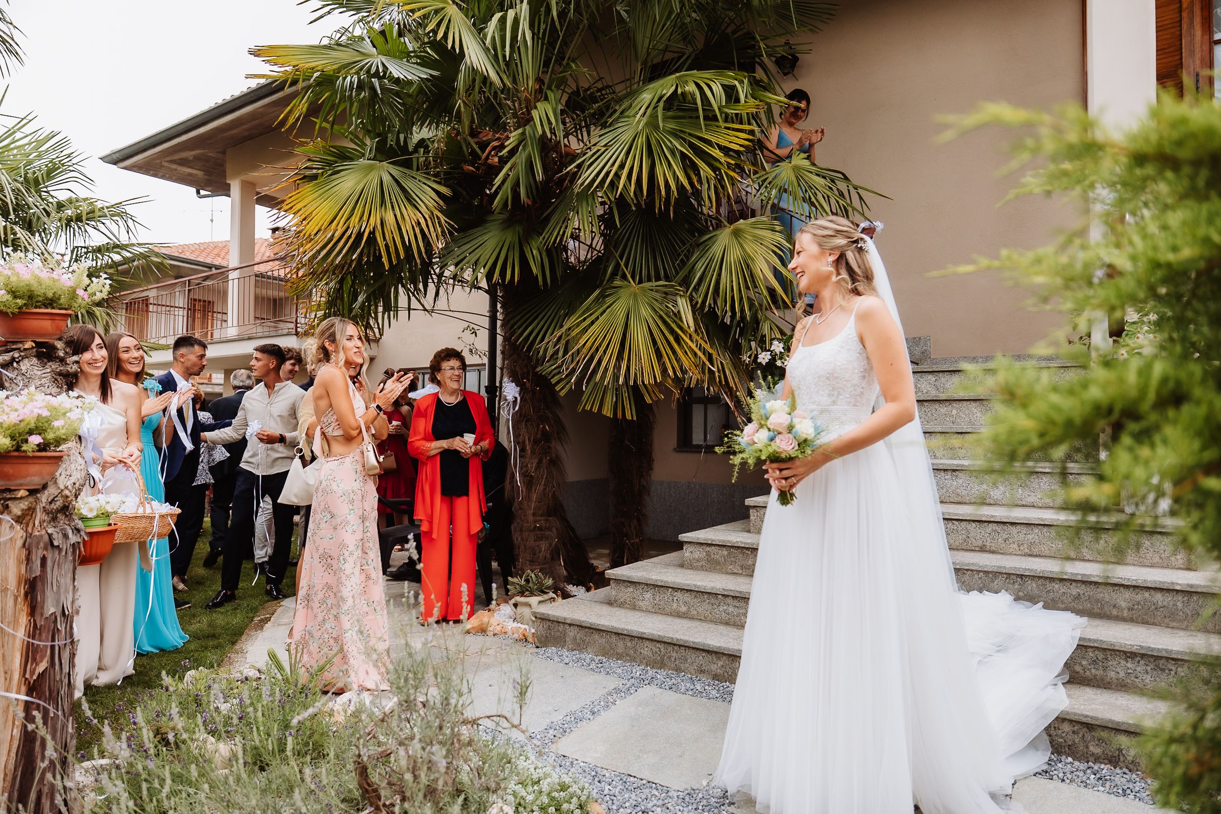 Wedding party members gather for photos on stairs at an outdoor venue with palm trees and greenery.
