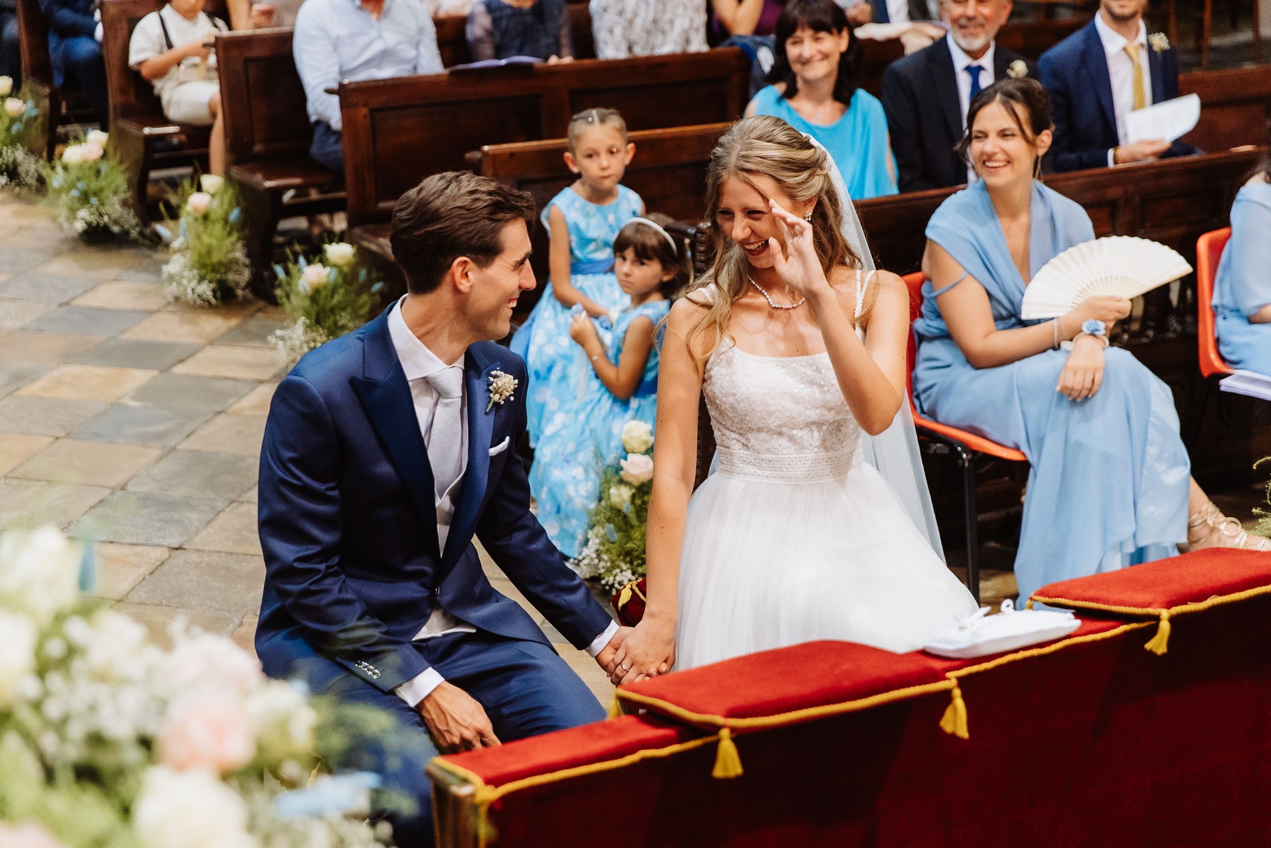 A couple smiles with joy as they ride in a red carriage on their wedding day, surrounded by loved ones.