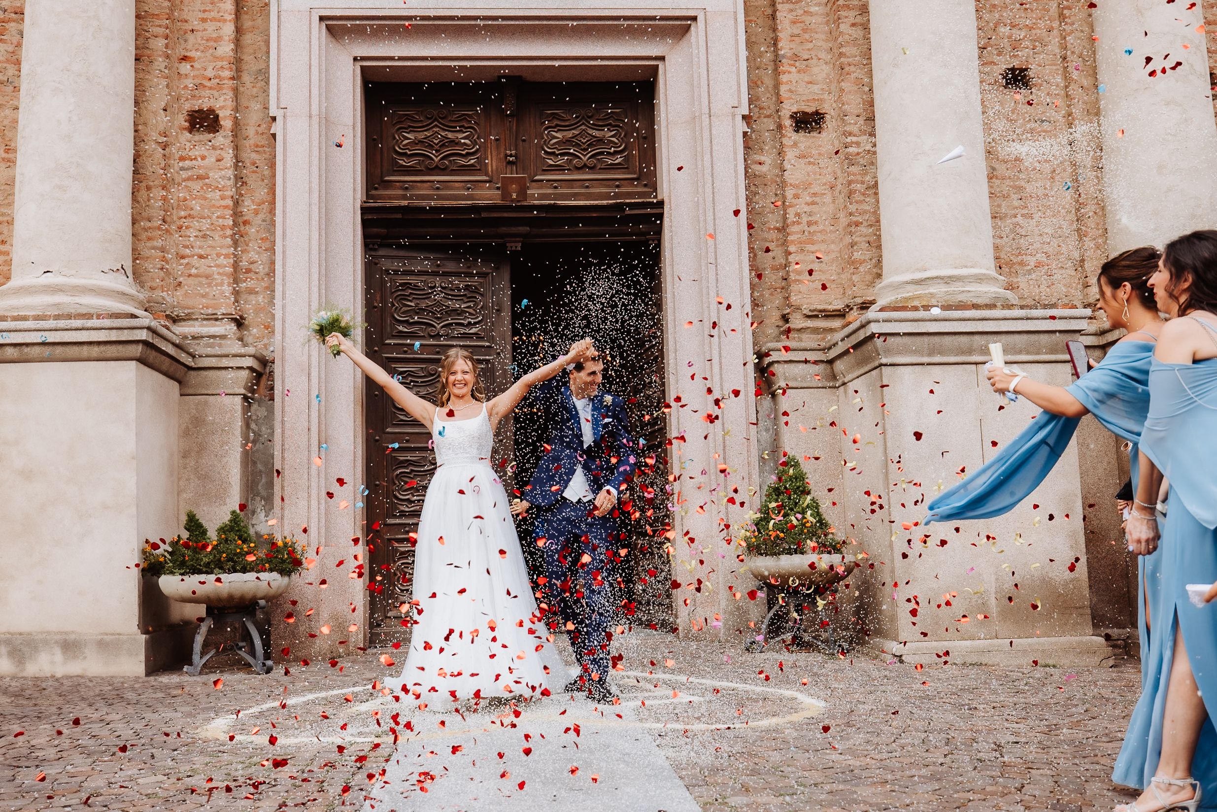 Wedding party celebrating outside historic stone church with pink and red flower petals tossed in the air.