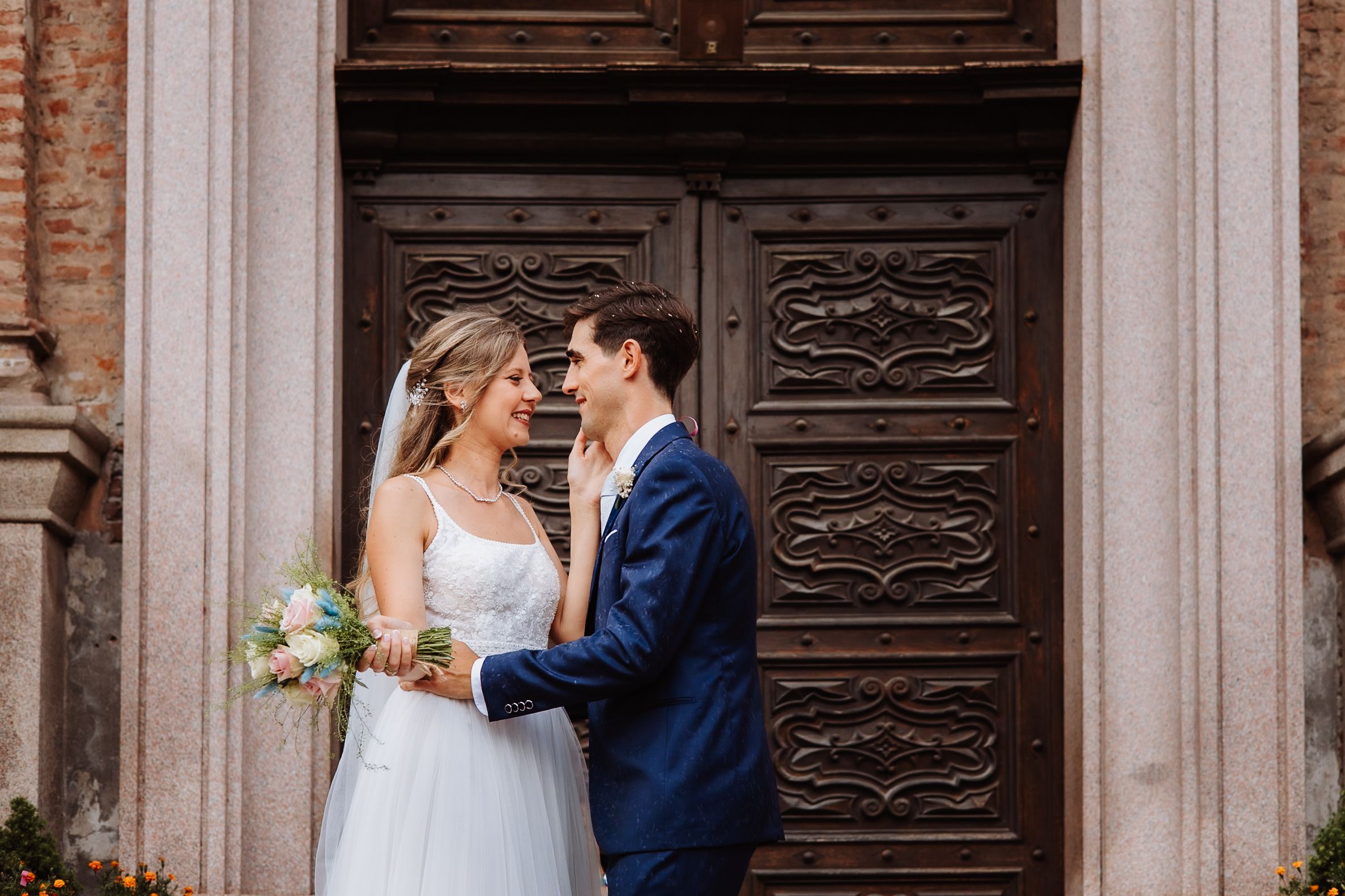 Elegant wedding couple poses near rustic wooden doors of historic stone building with ornate architectural details.