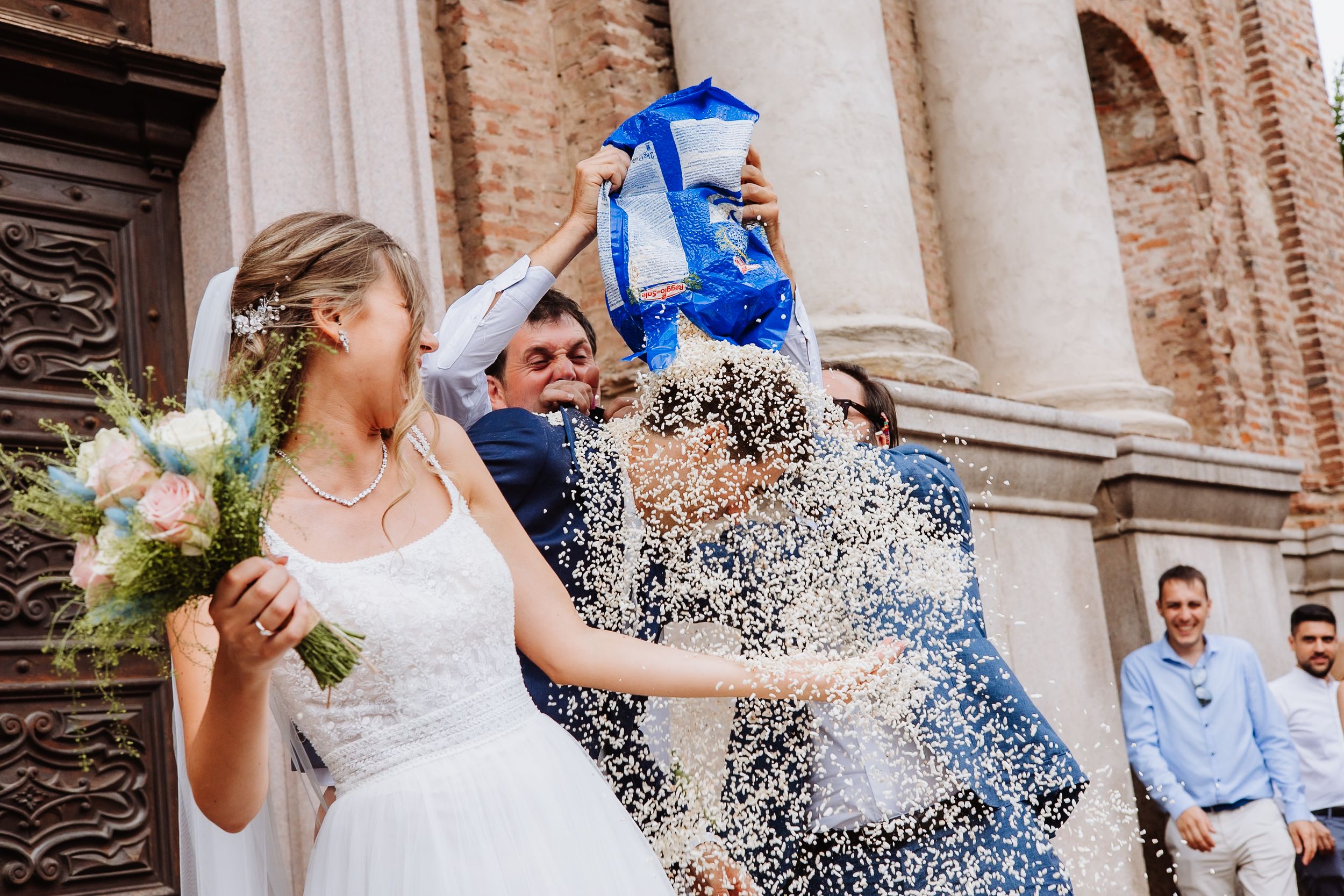Wedding guests throw handfuls of white confetti while celebrating the newlyweds outside a historic brick church.