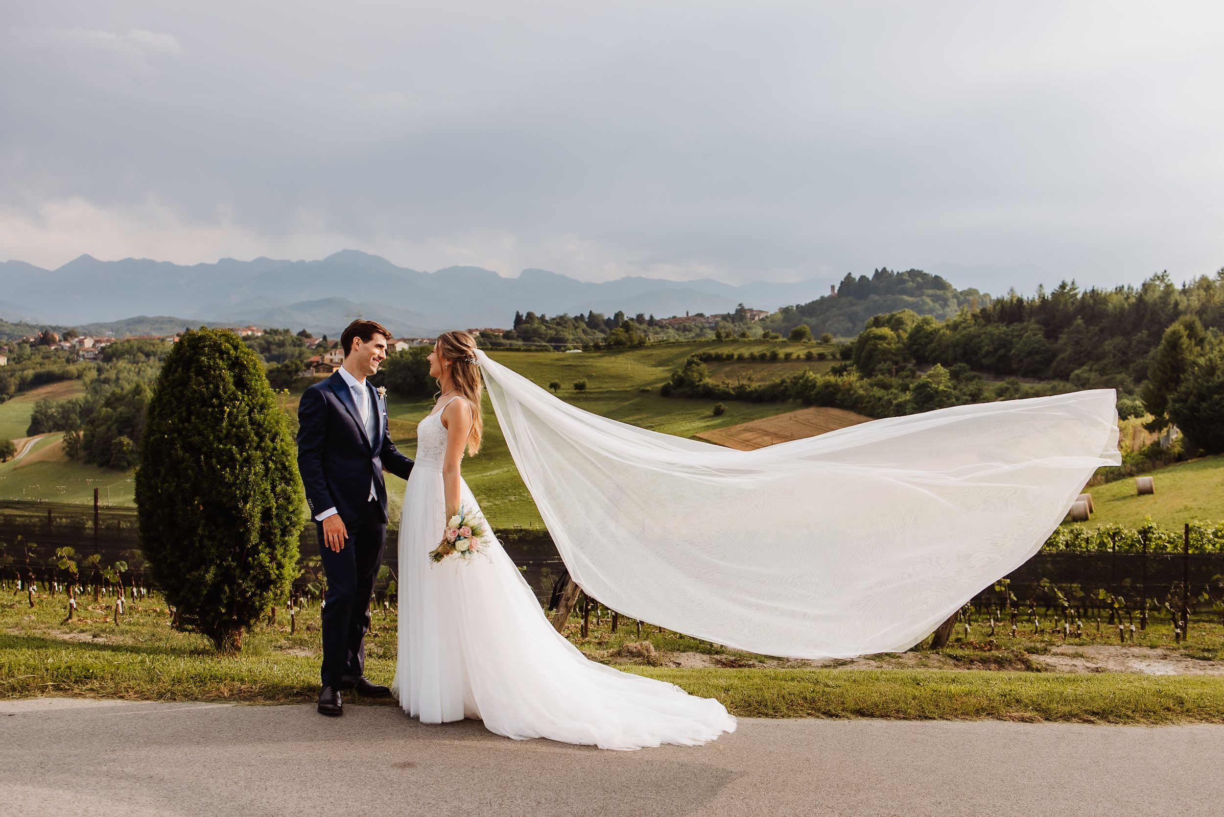 Wedding couple poses in scenic vineyard landscape with flowing white dress veil caught in wind against mountain backdrop.