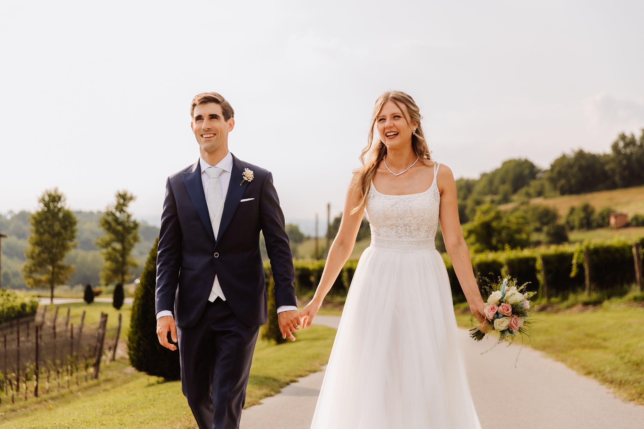 A couple walking hand in hand through a vineyard on their wedding day surrounded by lush countryside.