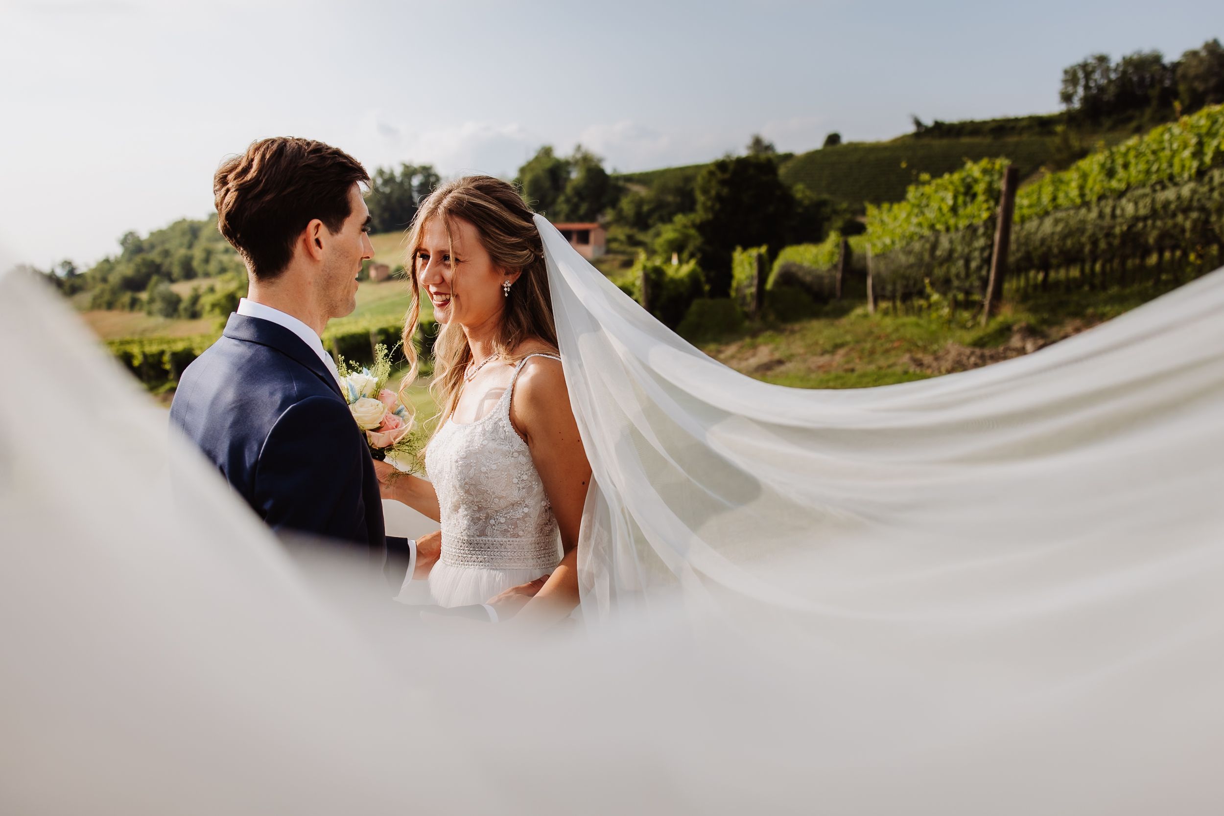 Wind catches the wedding veil as a couple embraces in a scenic vineyard setting.