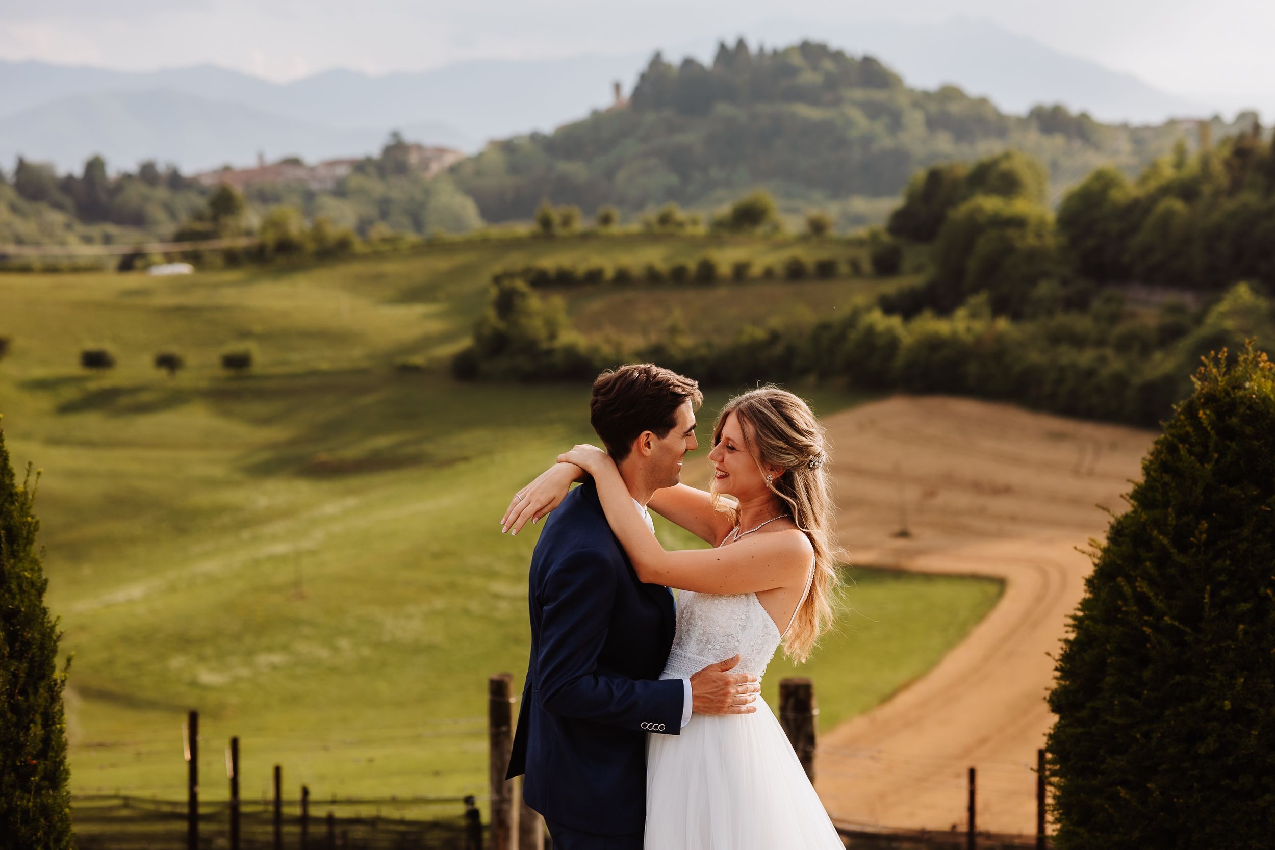 A romantic wedding couple embraces on rolling green hills overlooking a scenic Tuscan countryside landscape.