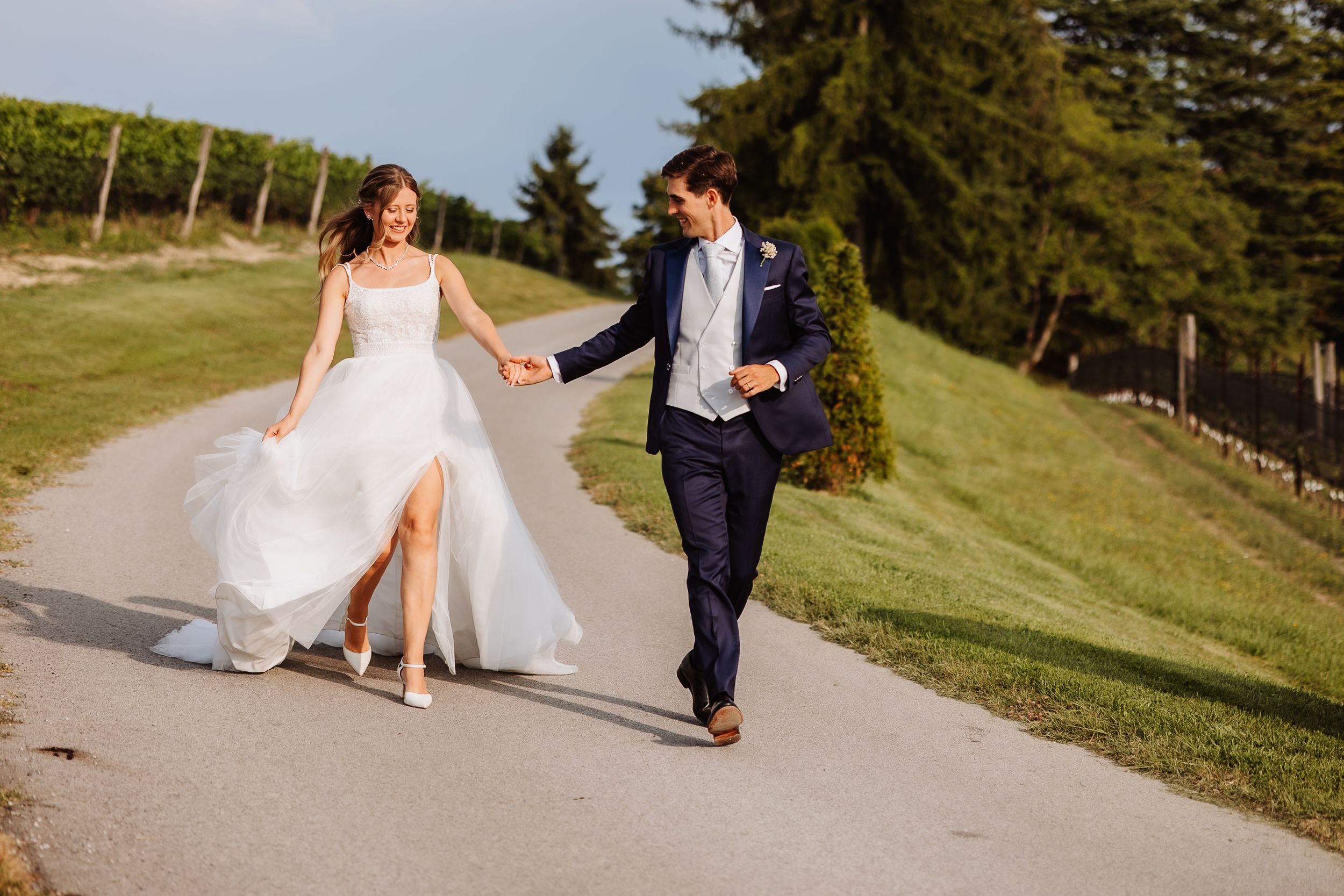 Couple holds hands walking across mountain path during outdoor wedding day photoshoot in nature setting.