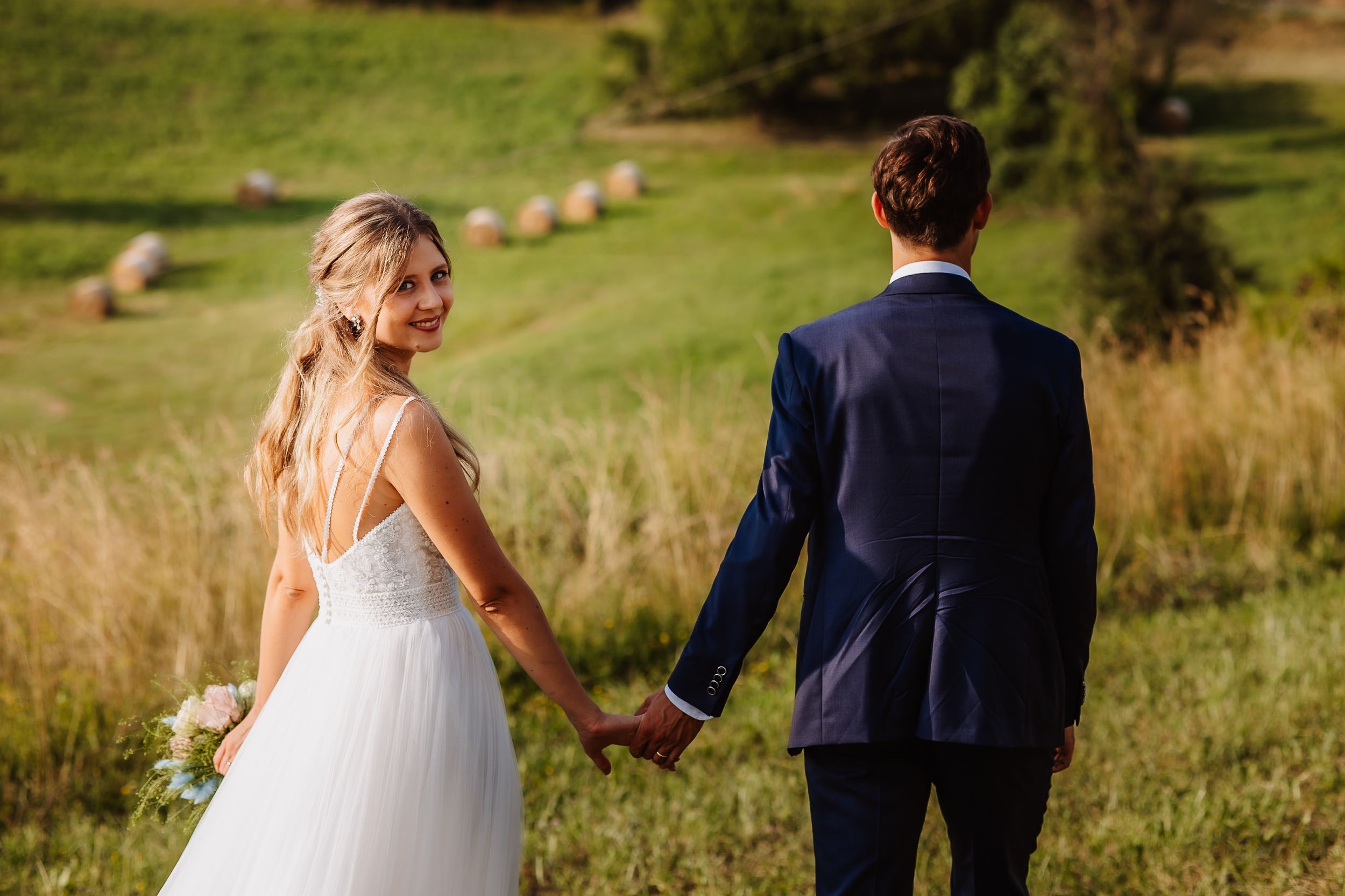 A couple in wedding attire walks across a lush green meadow on their special day.