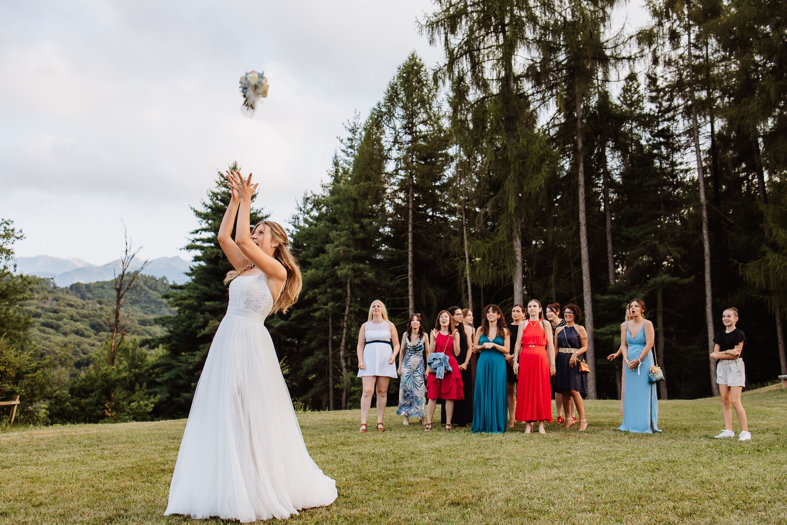 Scenic mountain wedding celebration with guests dressed in colorful attire gathered on a grassy hillside in the forest.