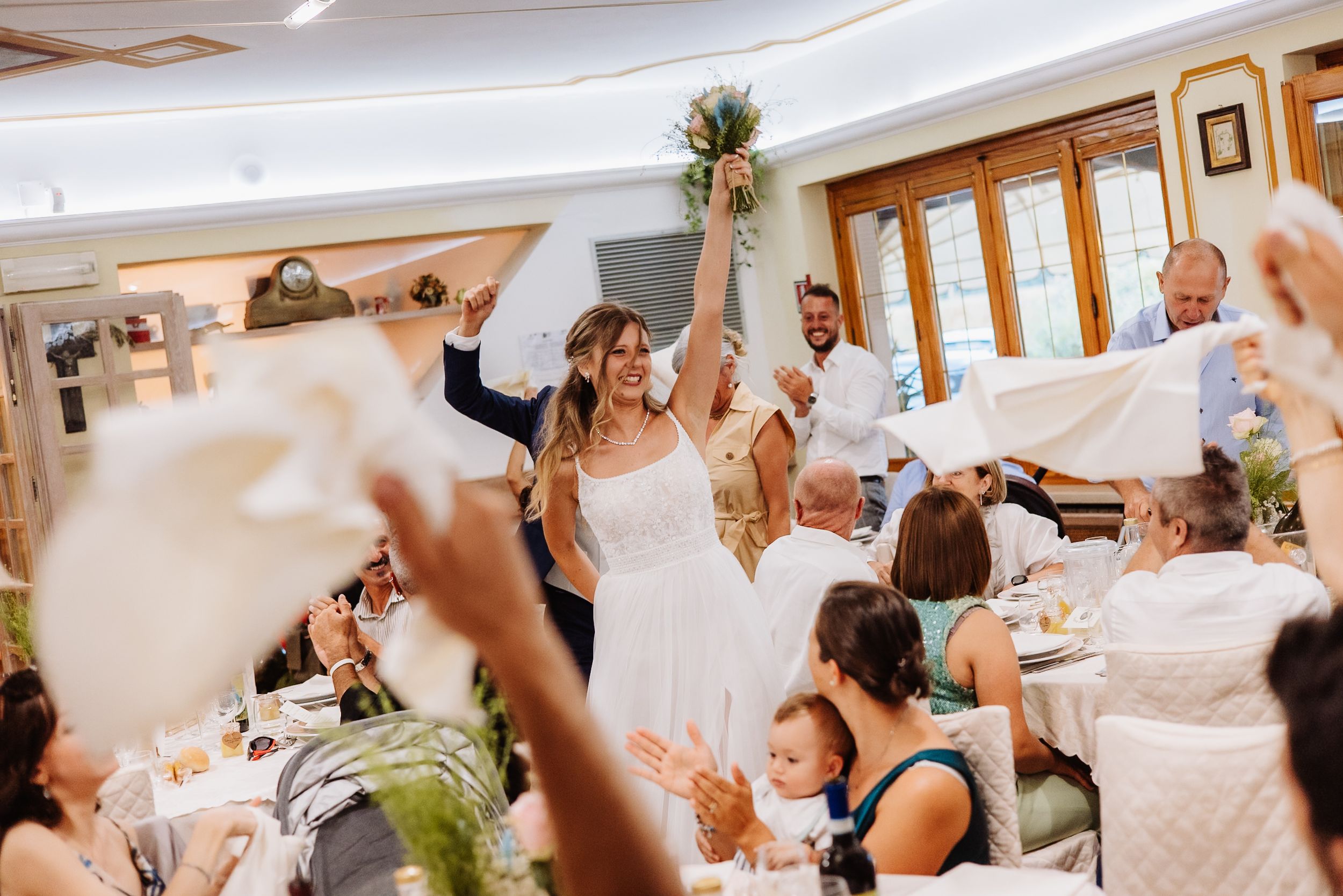 Wedding guests at a reception stand and wave white napkins during a celebratory dining hall moment.