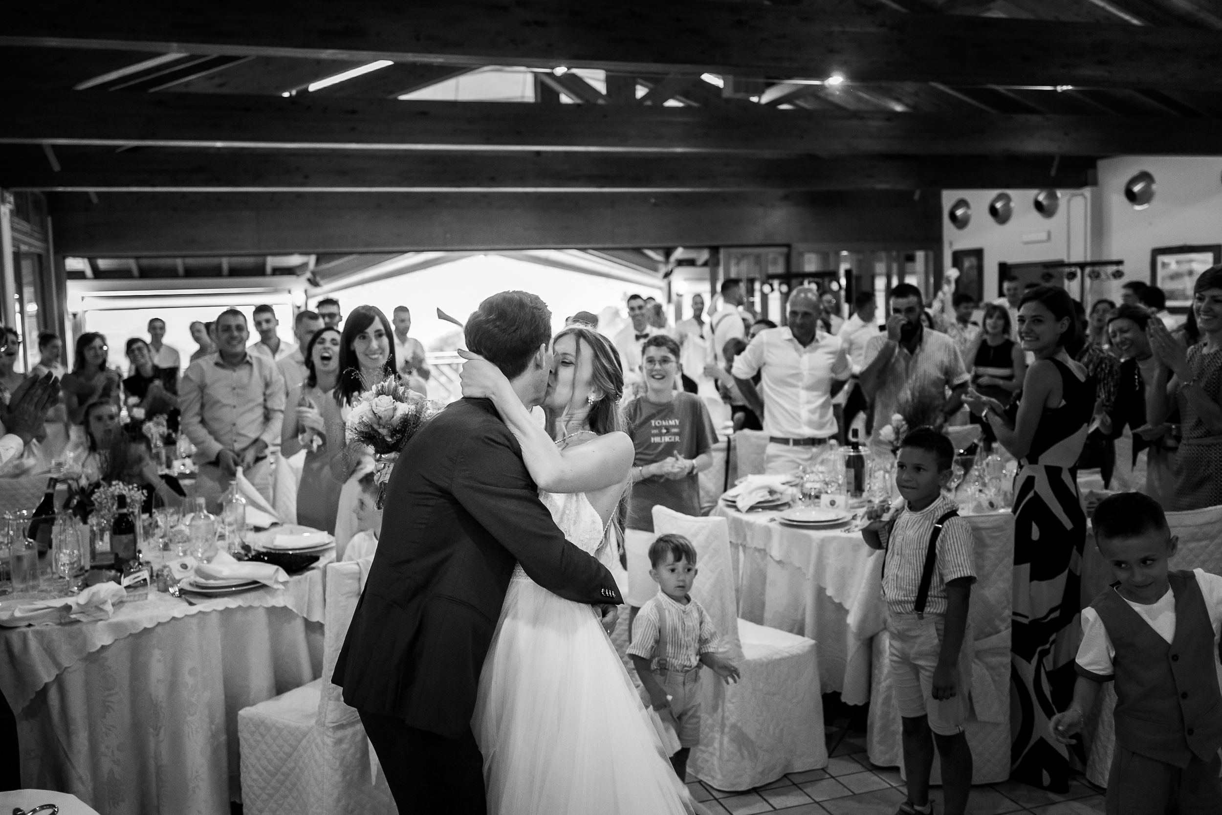 Wedding reception guests dance and celebrate on a monochrome dance floor under ambient lighting at an indoor venue.
