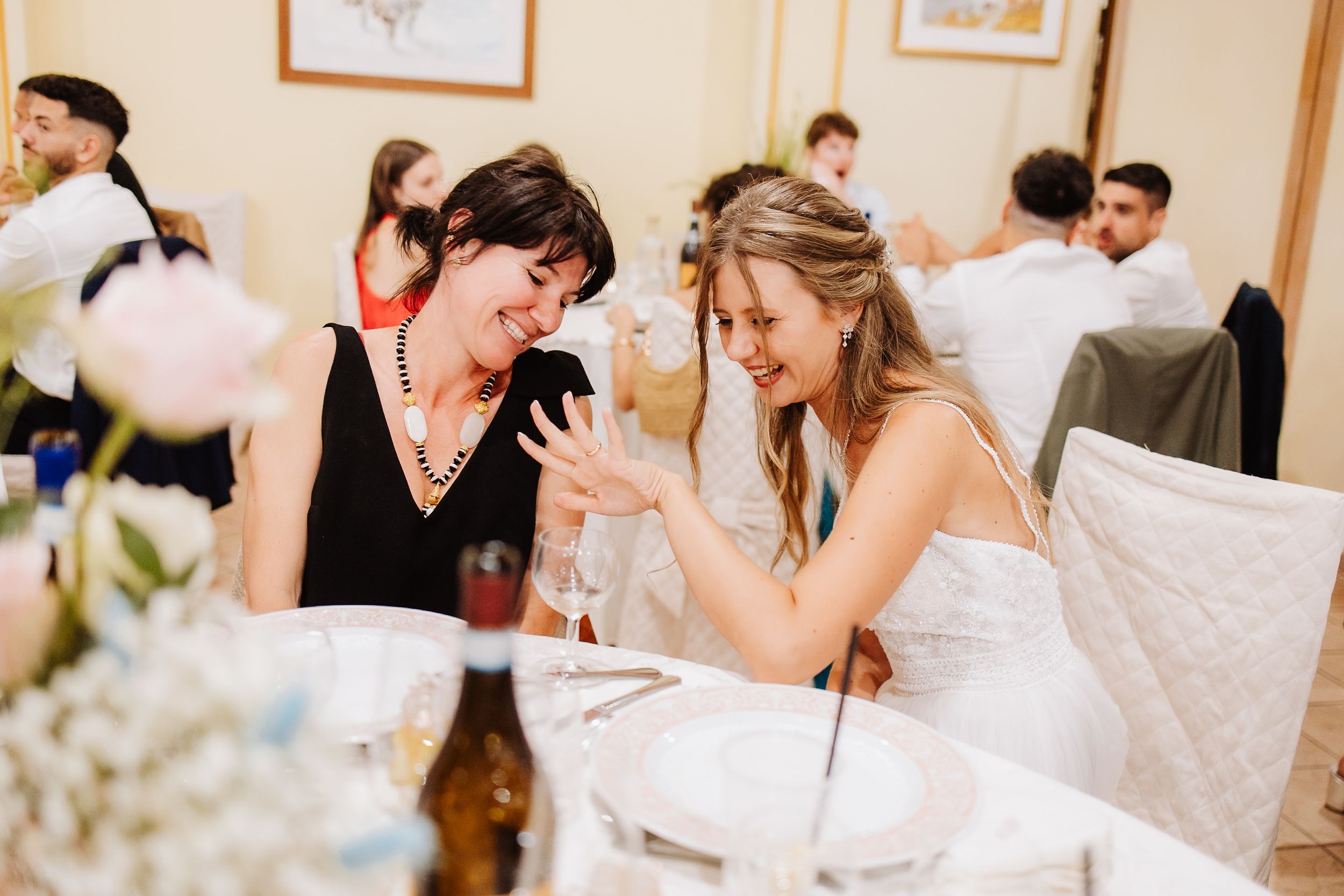 Two guests share an emotional moment at an elegant wedding reception with table decorations and other guests in background.