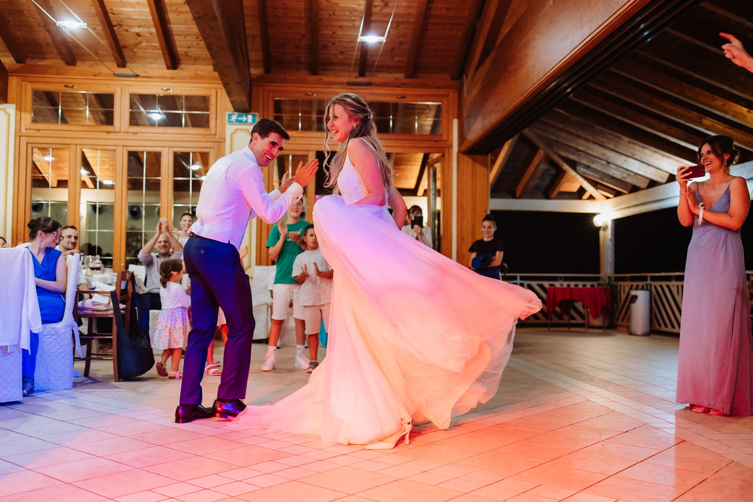Couple dances on illuminated floor during wedding reception in rustic wood-beamed venue with pink lighting.