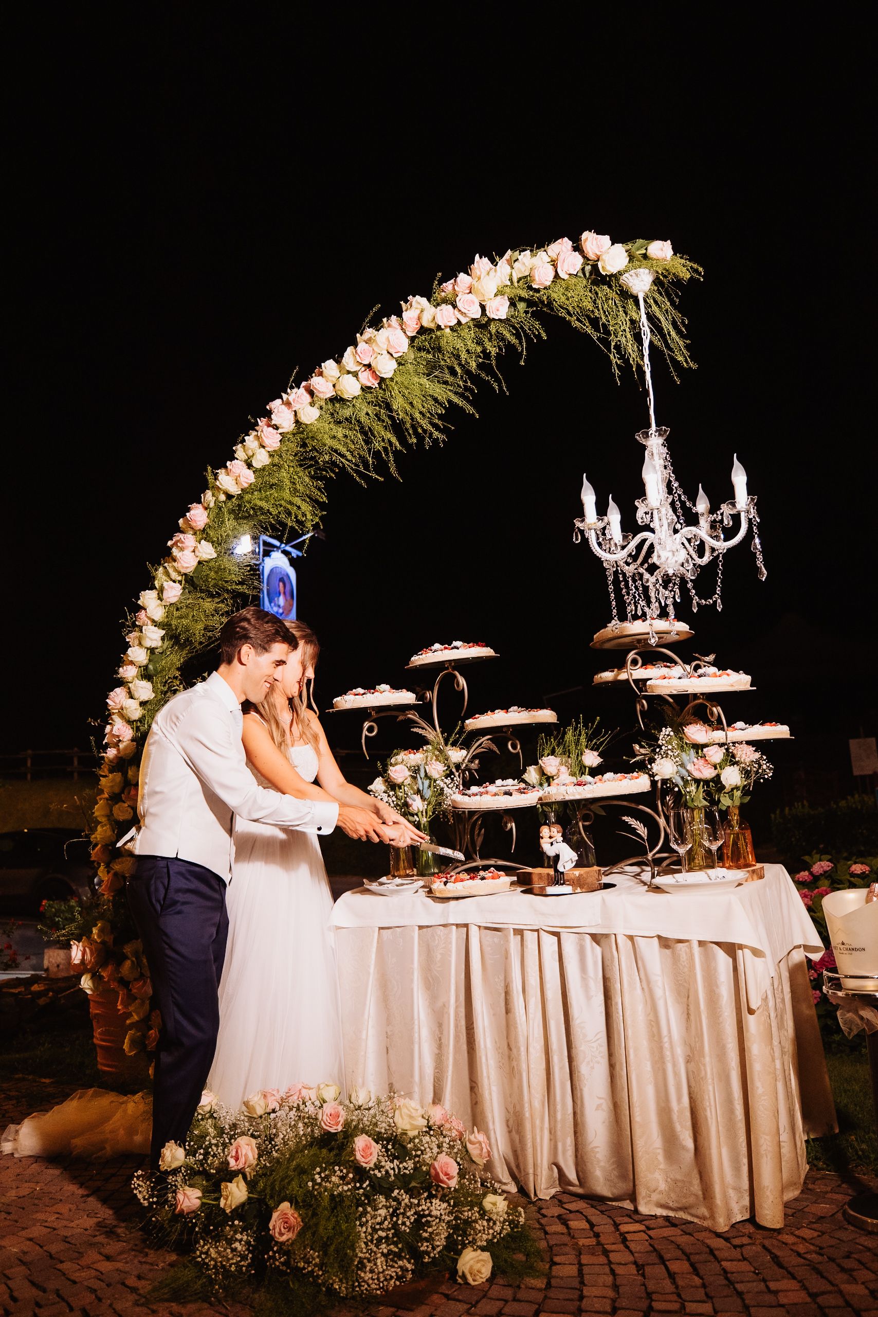 Couple cuts their wedding cake together under an elegant floral arch at an outdoor evening reception.