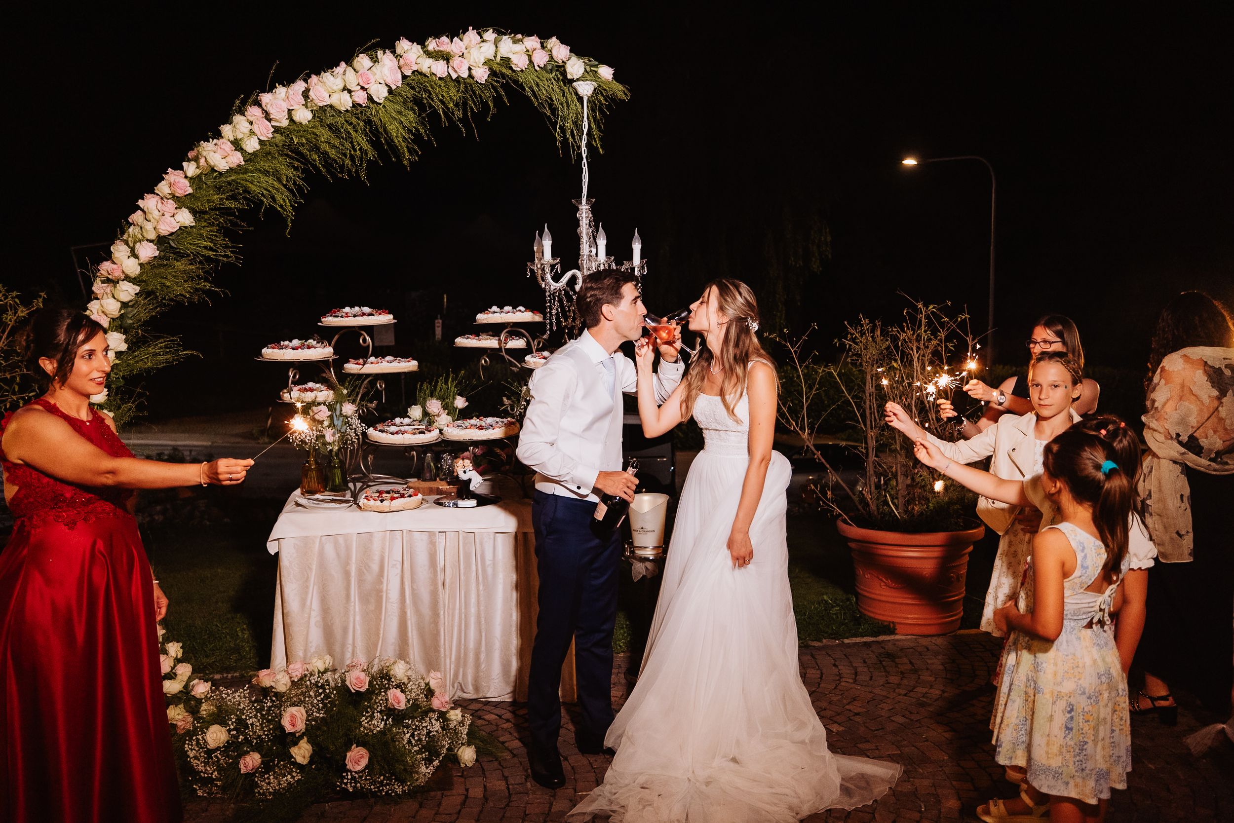 A romantic wedding celebration at night with a floral arch and elegant dessert display table.