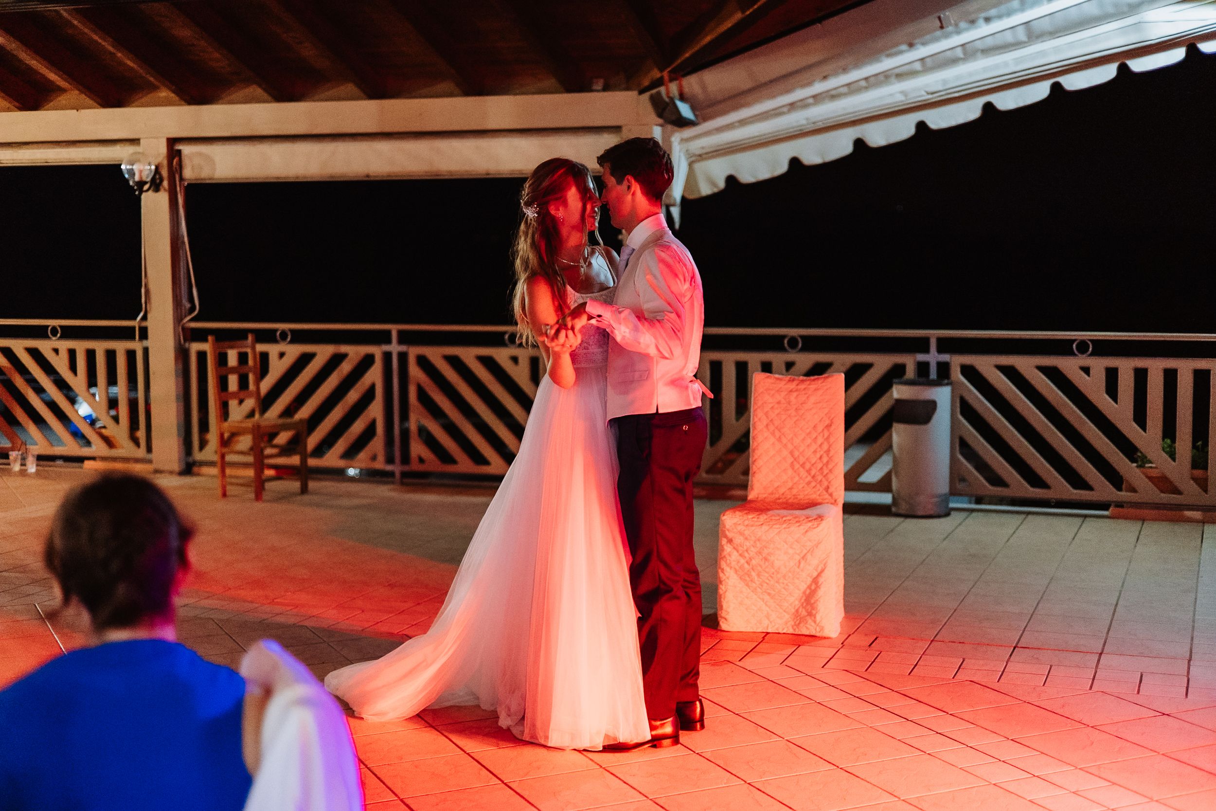 A couple shares their first dance at a wedding reception on an outdoor patio illuminated by red and blue lighting.