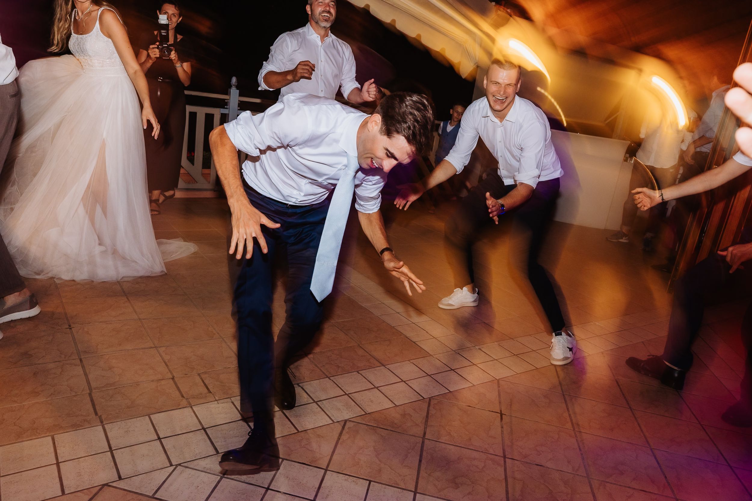 Guests dancing together during a wedding reception on a tiled dance floor under warm amber lighting.
