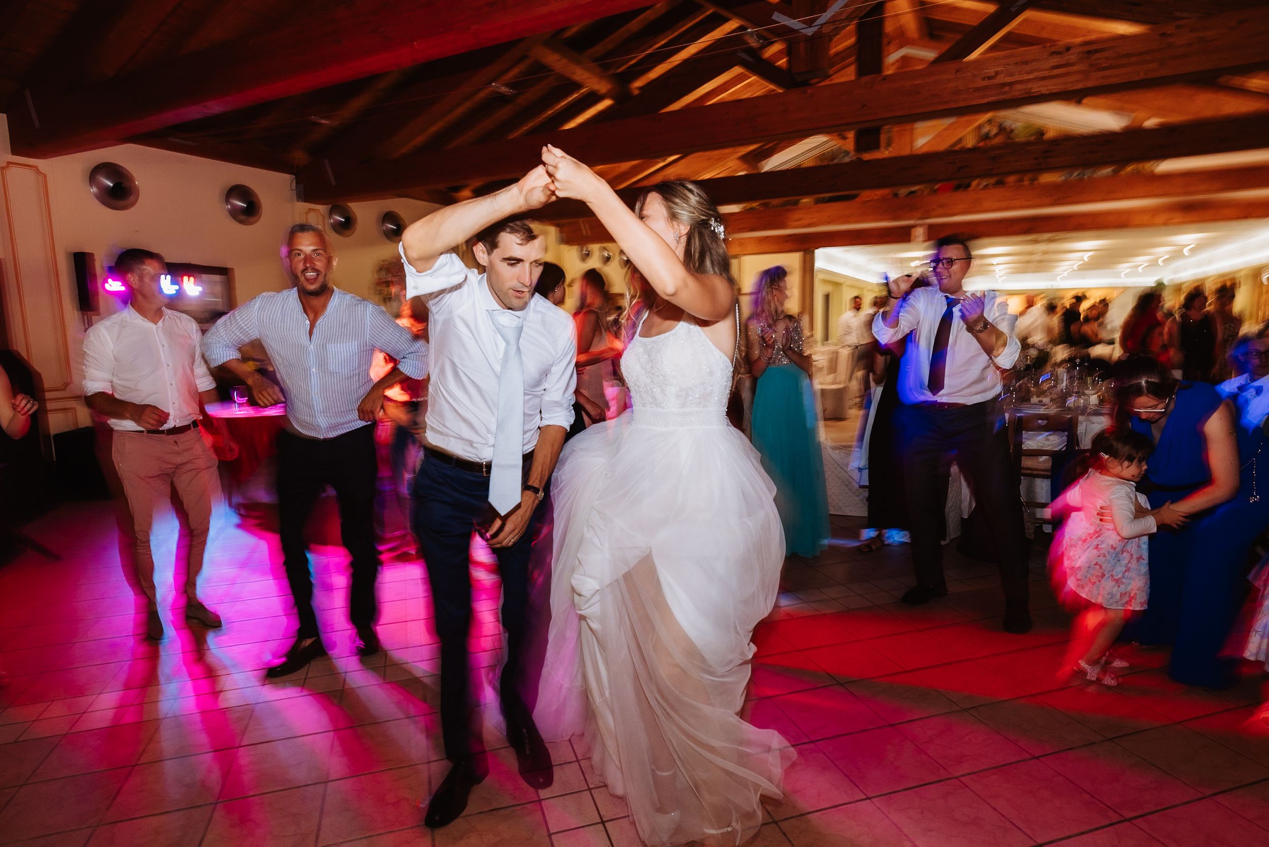 Wedding guests dancing and celebrating on an illuminated dance floor with pink lighting at a reception venue.