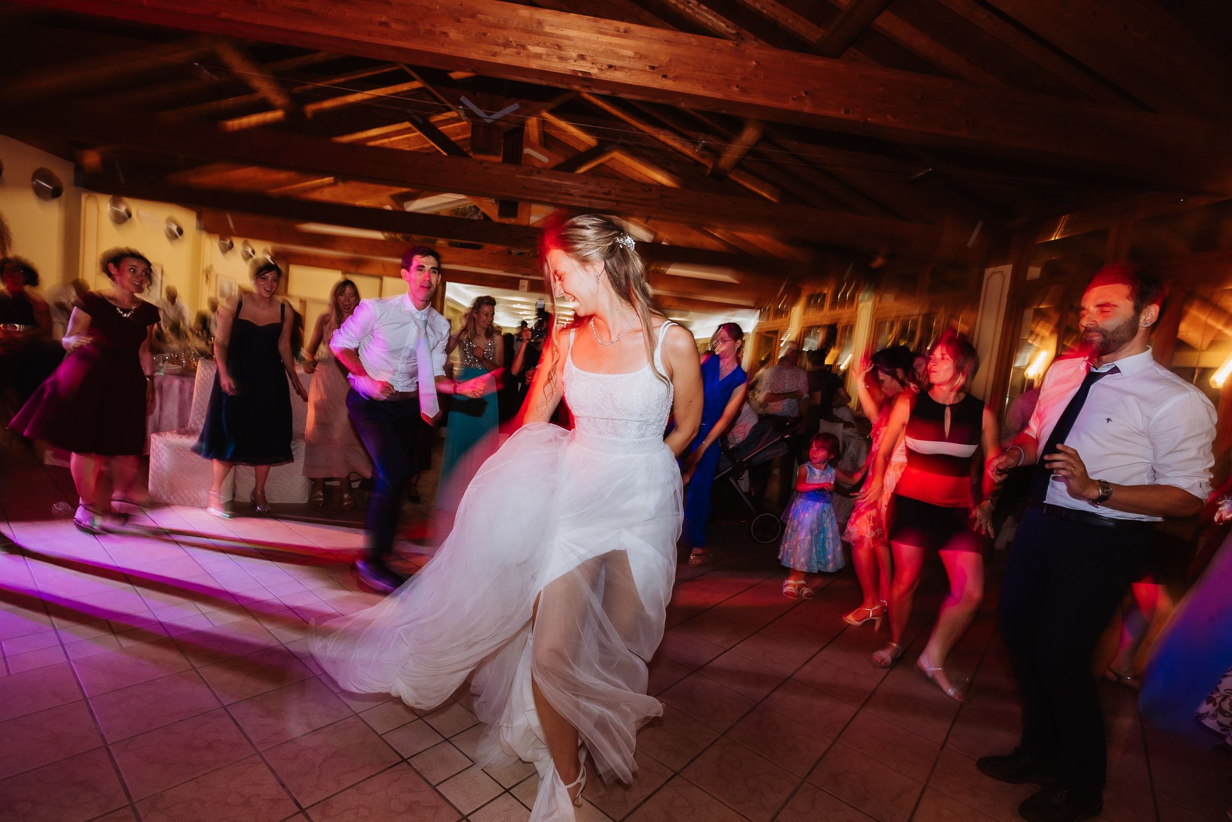Wedding guests dance and celebrate on a rustic wooden dance floor under warm lighting at an indoor reception.