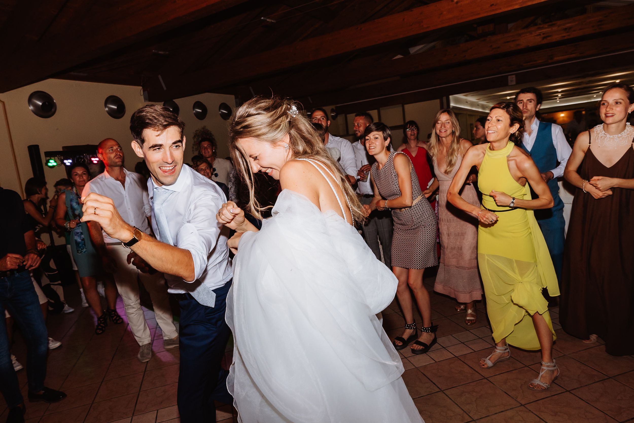 Couples dance together during a lively wedding reception in a dimly lit rustic venue with wooden beams.